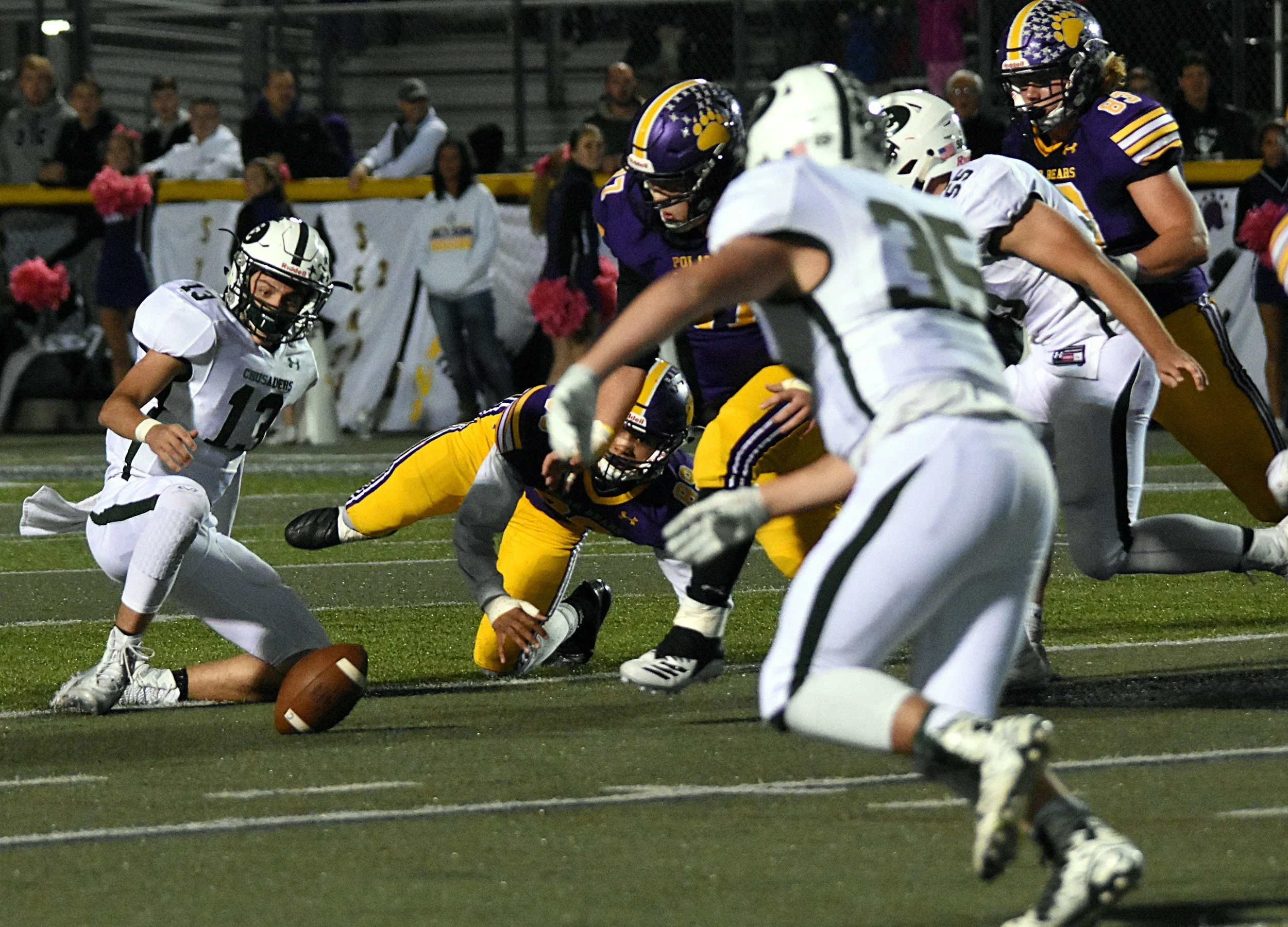 Football players in purple and white uniforms compete for control of the ball during a game at night, with spectators watching in the background.