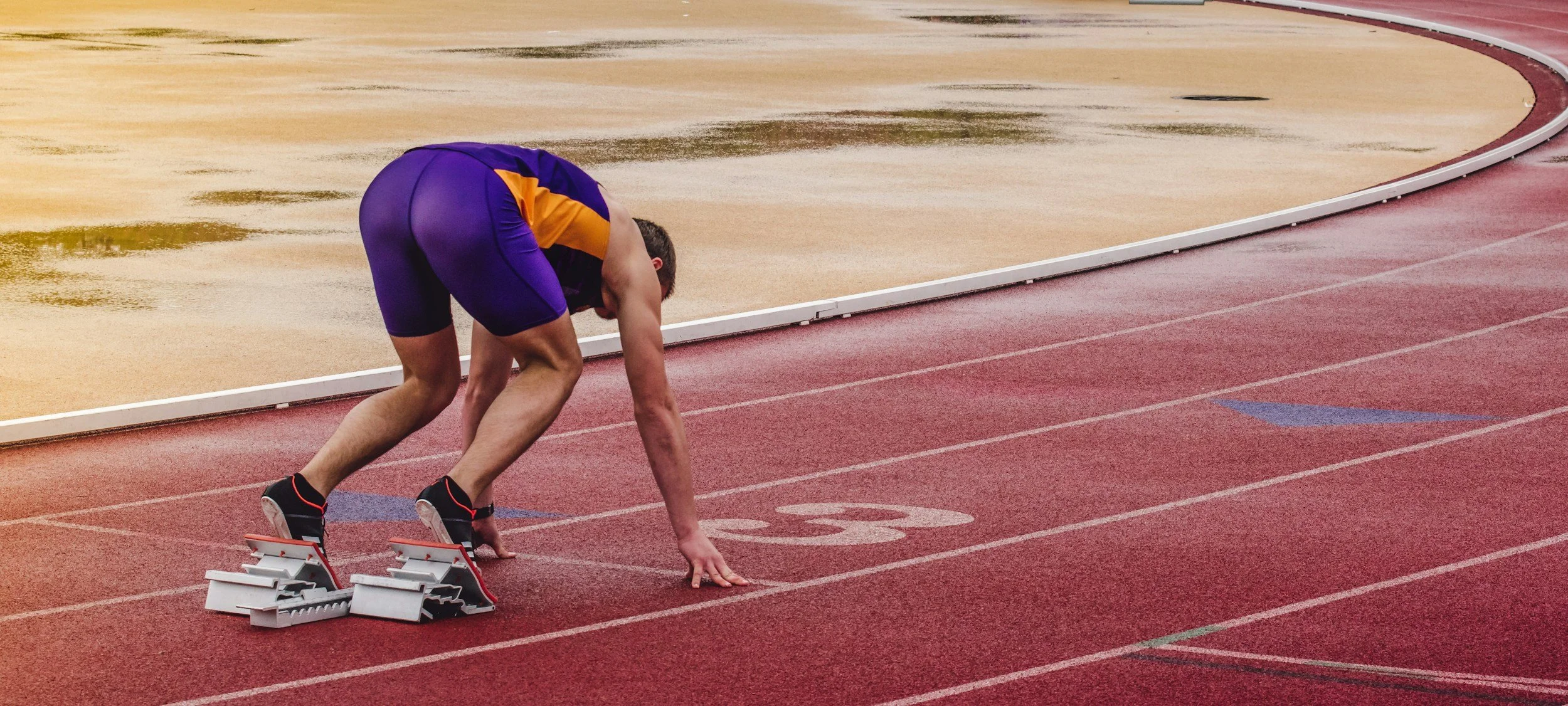 A male sprinter in a starting position on a track at an athletics stadium, wearing a purple and yellow racing uniform and black running shoes, with starting blocks set in front of him.