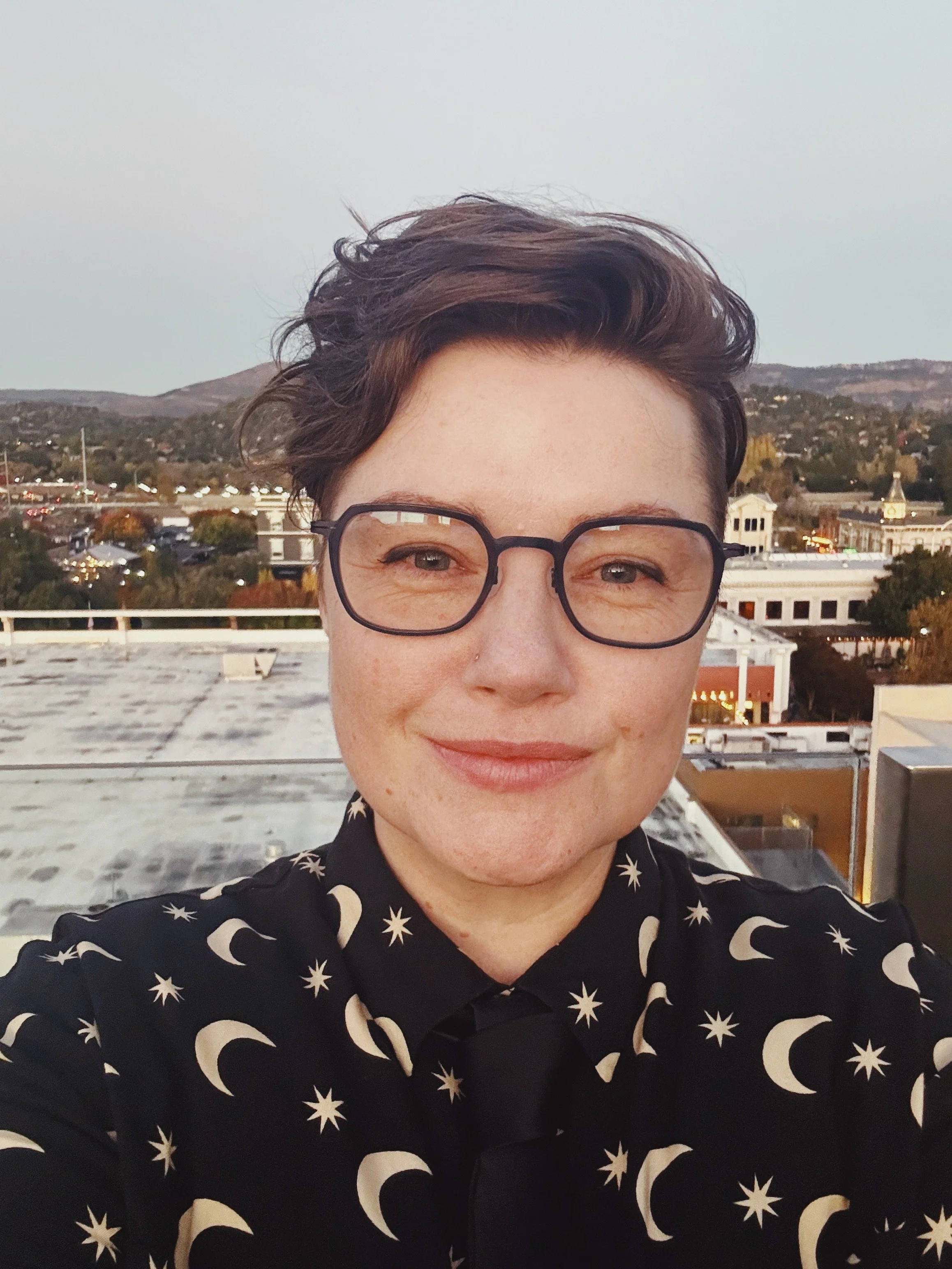 Tal smiles gently at the camera: they are a white person with square-ish glasses and short, wavy  light brown hair, wearing a black shirt decorated with stars and moons.