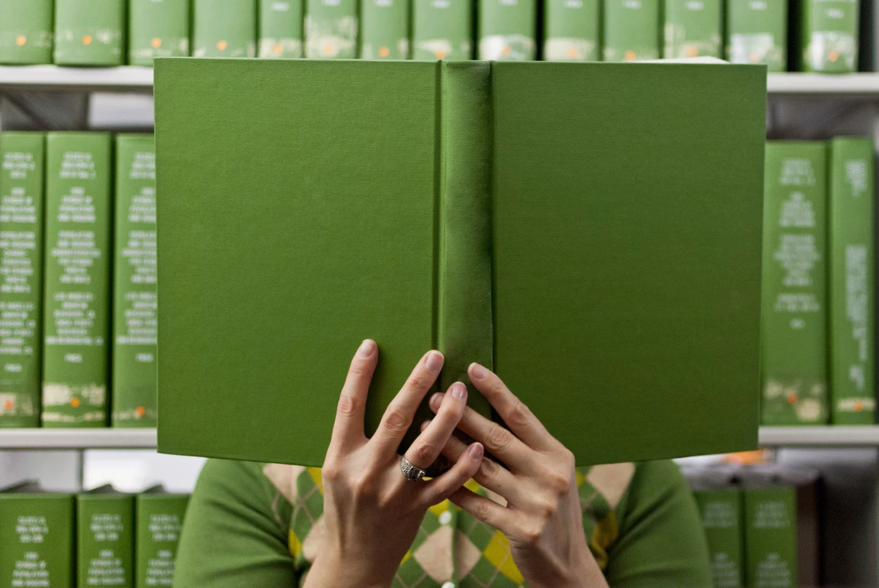 A green book being held up by a person with white skin. The book obscures their face.