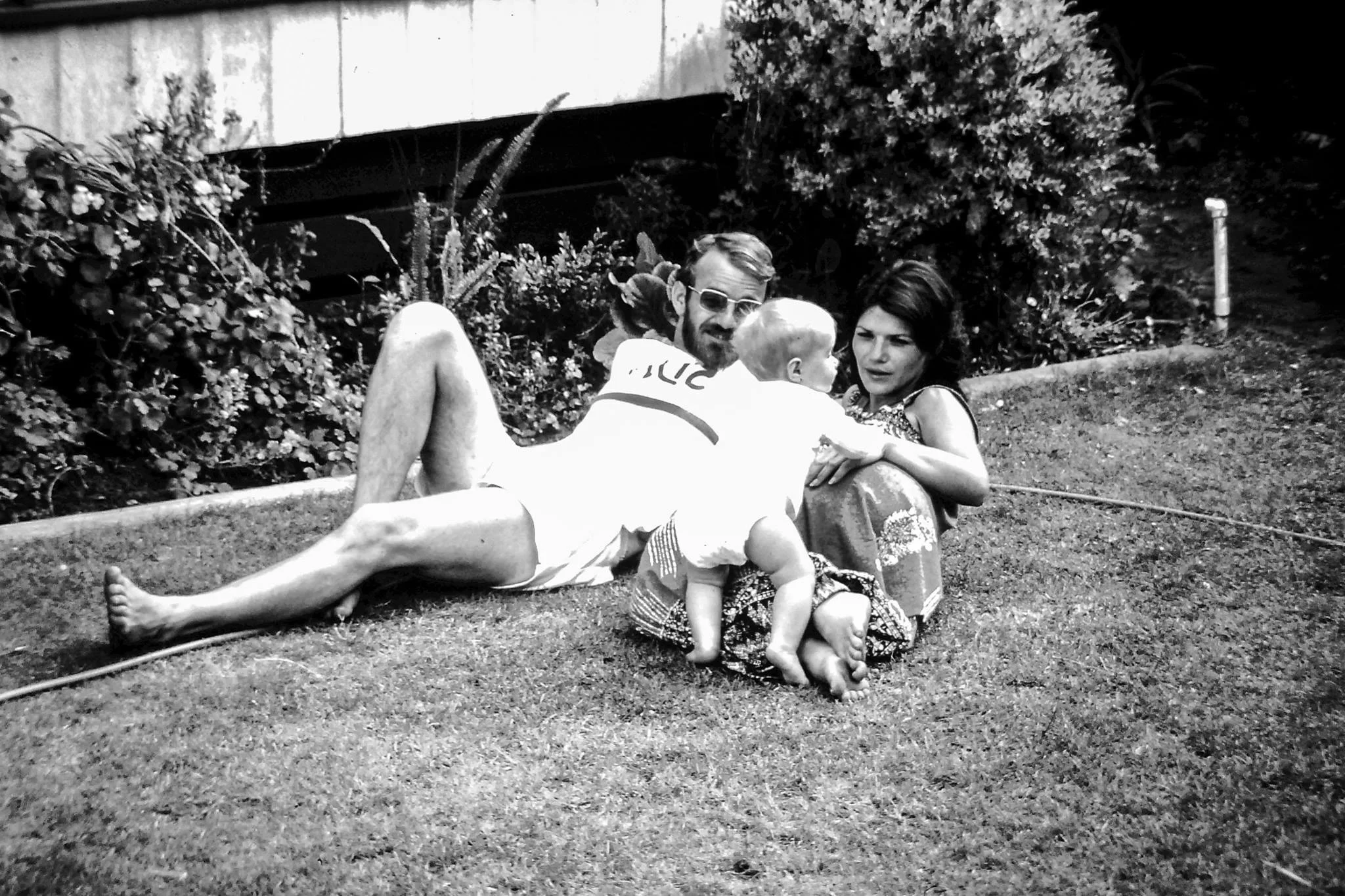 In this black-and-white photo, two adults recline on a lawn with their baby in the late 1970s. They're dressed for summer, and are looking at the baby as she leans on her mother's legs.
