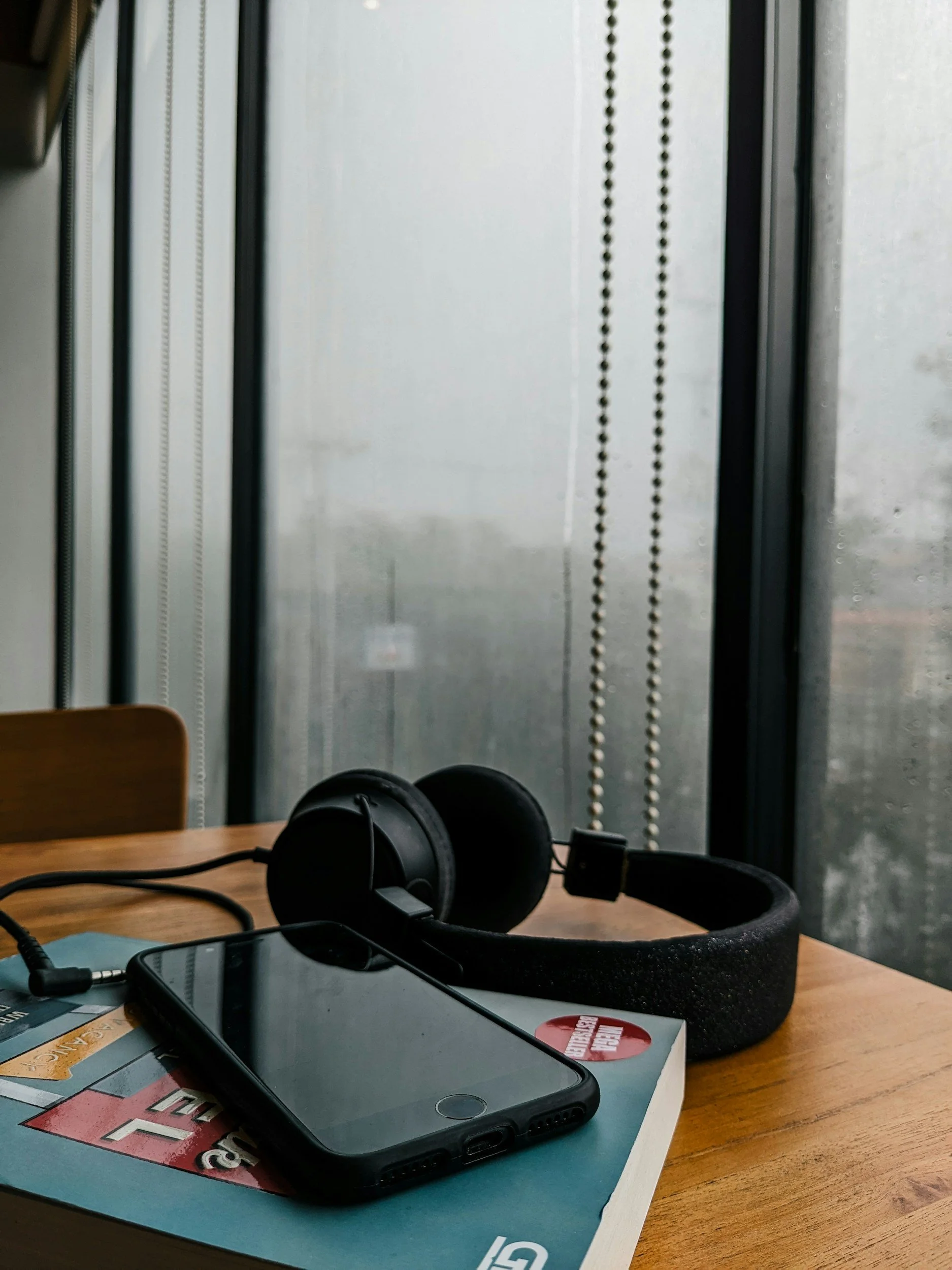 Book Edits, Proposals, Ghostwriting, Podcast and Audiobook Narration and Production. Image is of a pair of black headphones, a smartphone, and a hipster magazine on a wooden table near a window with rain droplets.