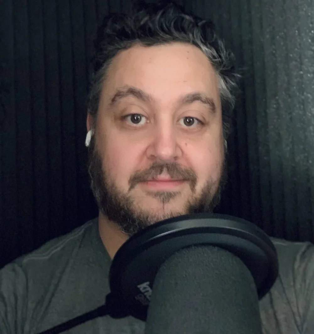 Andrew Weber of APW Author Services. He has dark hair and a beard and is wearing earbuds, speaking into a professional microphone in a recording studio with dark soundproofing panels.