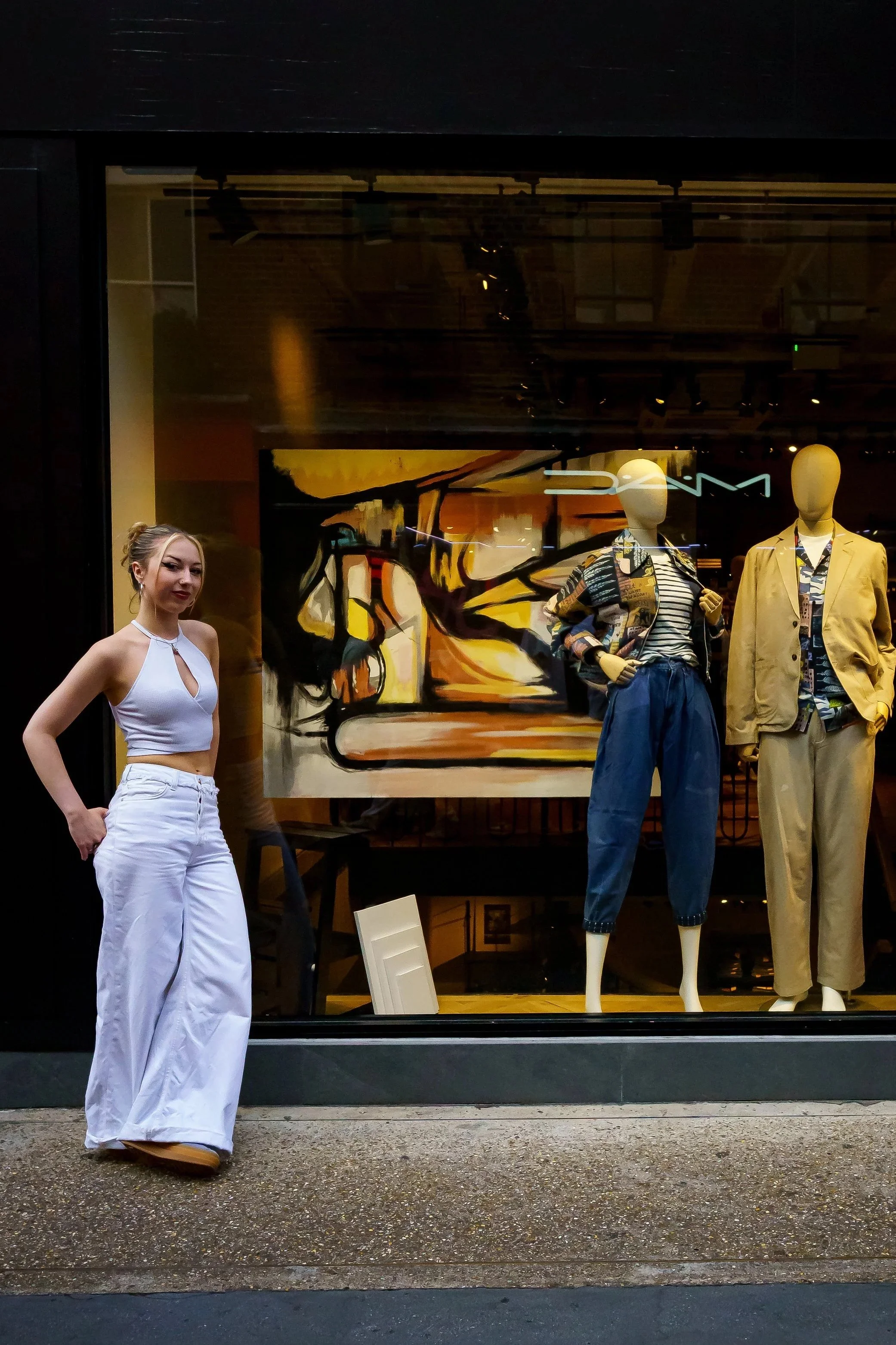 A woman in white pants and a halter top standing outside in front of a store window display with mannequins dressed in casual clothing and a large abstract painting in the background.