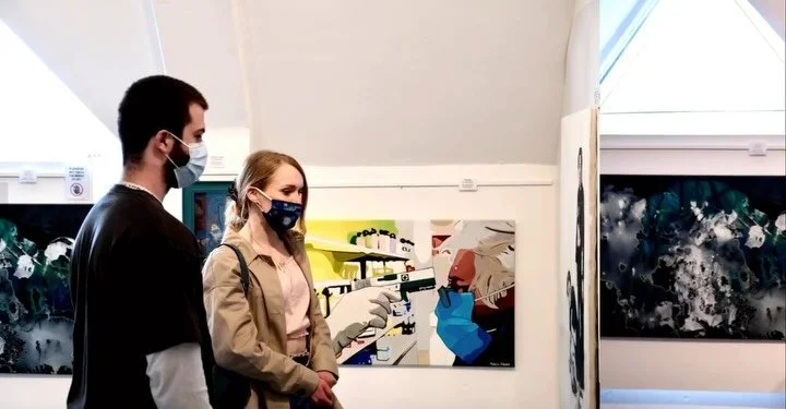 Two people, a man and a woman, wearing masks, standing in an art gallery looking at paintings.