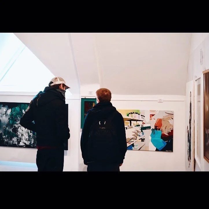 Two men look at artwork in an art gallery with white walls and a sloped ceiling.