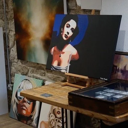 Art gallery with paintings, including a stylized portrait of a woman and abstract artwork, displayed on easels and leaning against a brick wall.