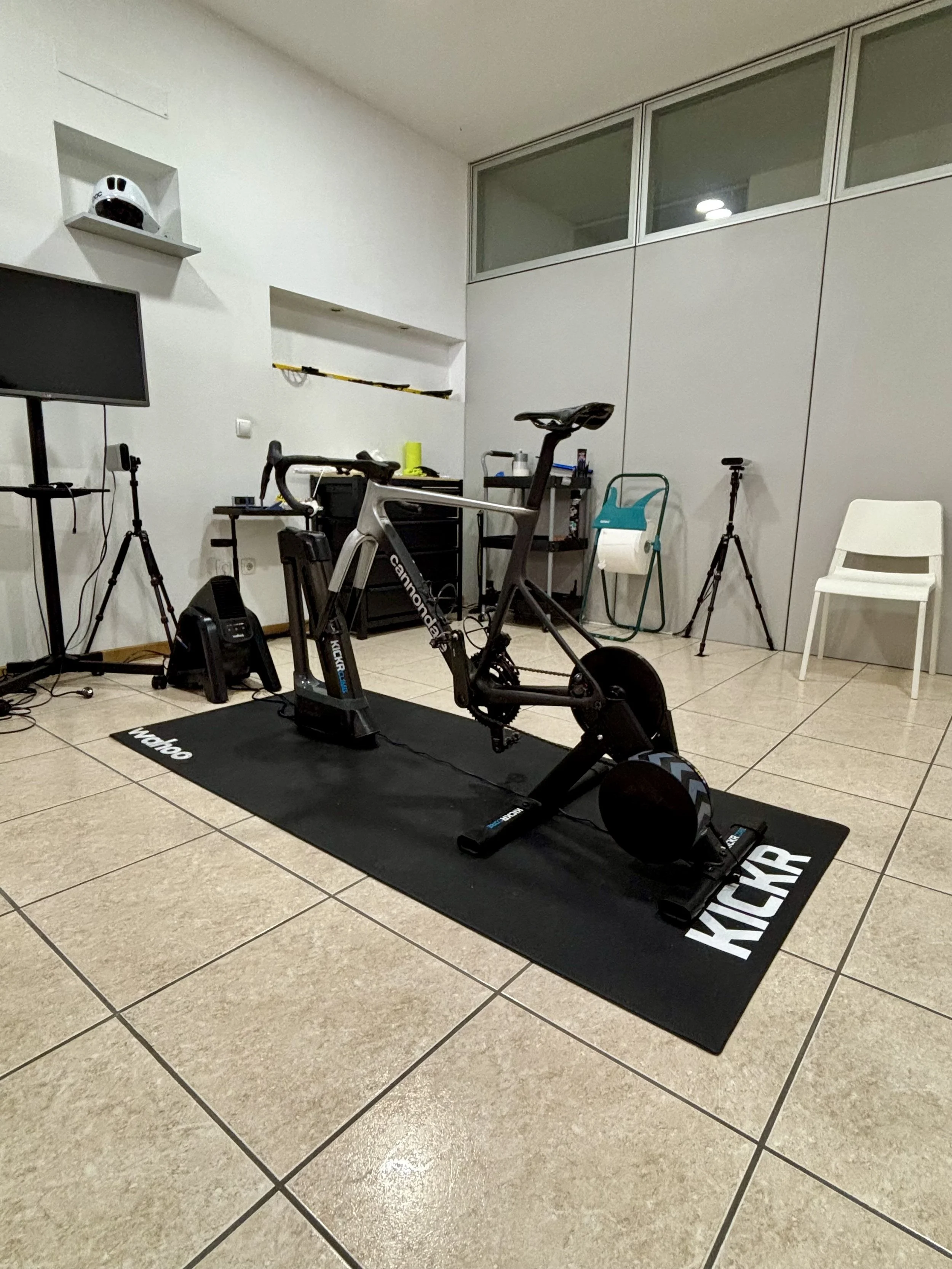 Sala com um cic kit, uma televisão, uma cadeira branca, uma cadeira azul, tripés, uma prancha de patinação, uma lousa preta com a palavra 'KICKR'.