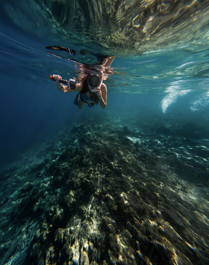 A person scuba diving underwater surrounded by rocks and coral, holding a camera.