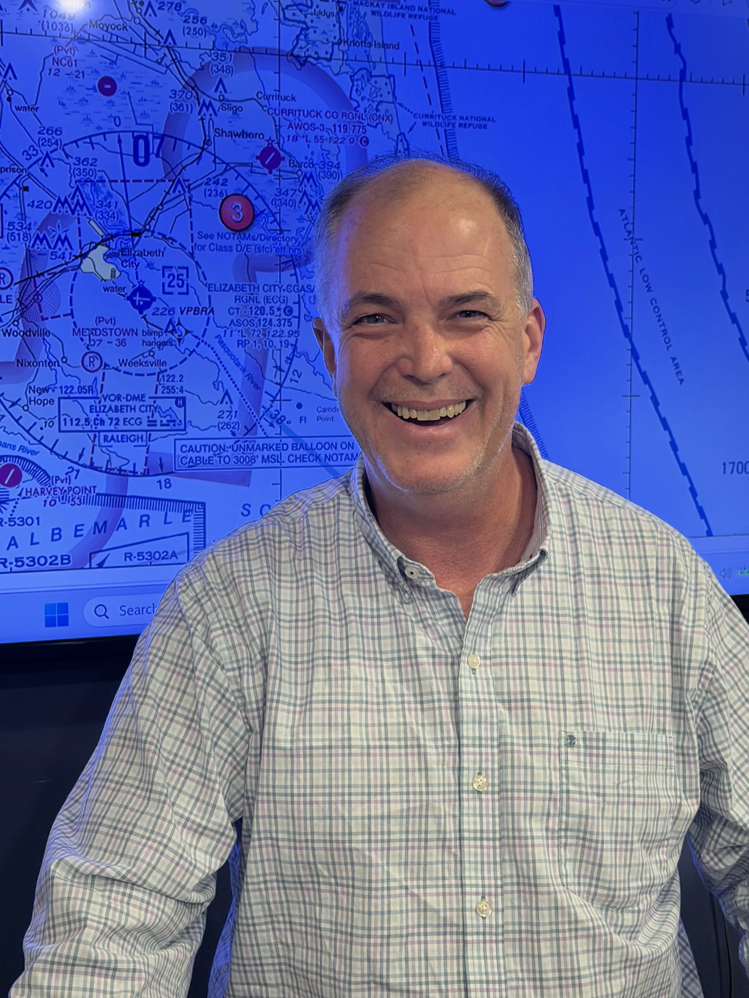 A smiling man wearing a checkered shirt standing in front of a blue screen displaying an aviation chart or map.