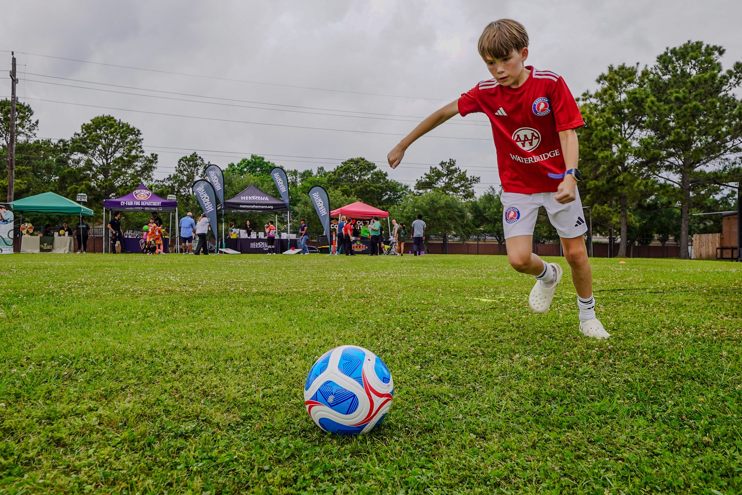 YMCA soccer player in a red soccer jersey and white shorts is about to kick a blue and white soccer ball on a grassy field during an outdoor event with tents and people in the background.