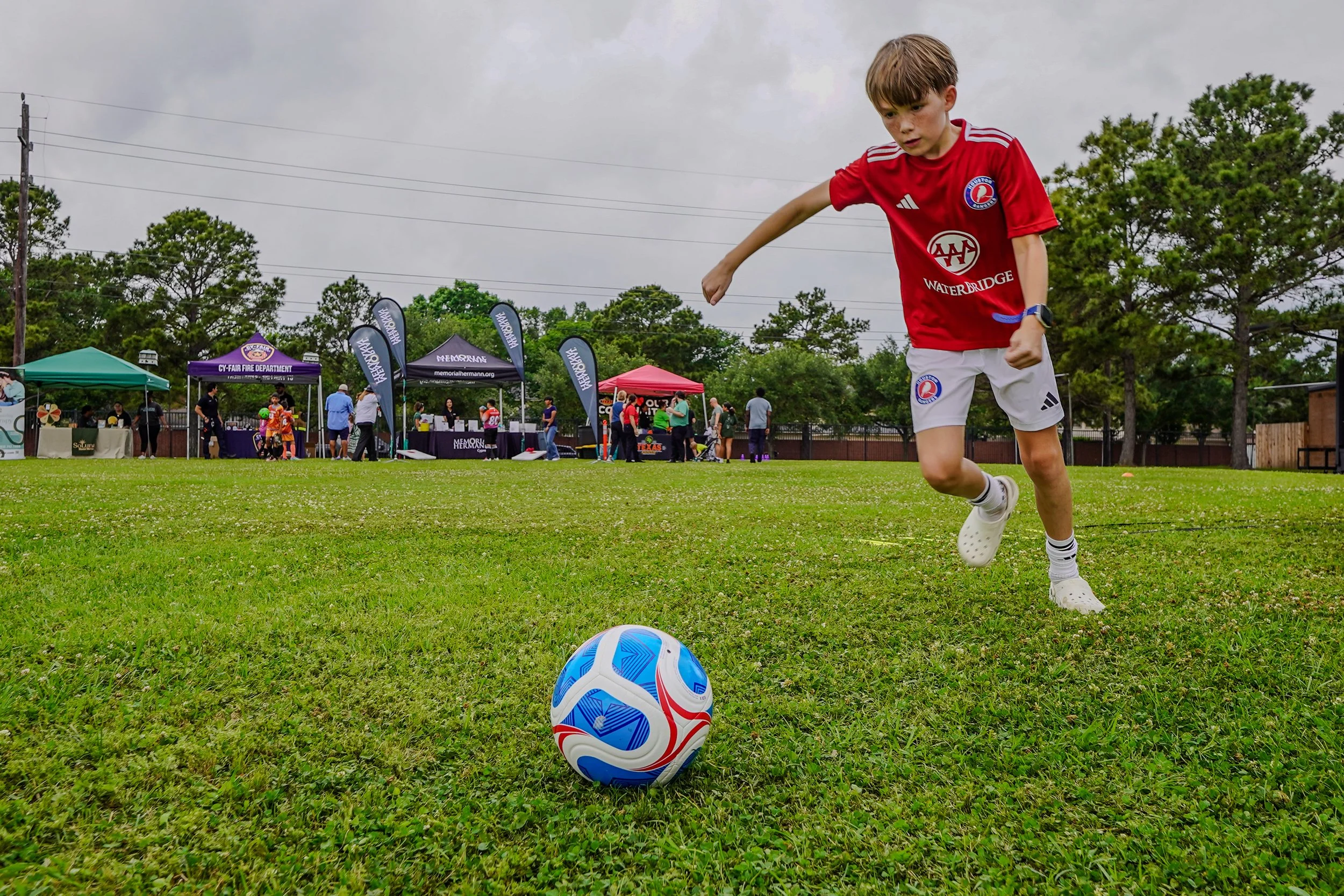 YMCA soccer player in a red soccer jersey and white shorts is about to kick a blue and white soccer ball on a grassy field during an outdoor event with tents and people in the background.