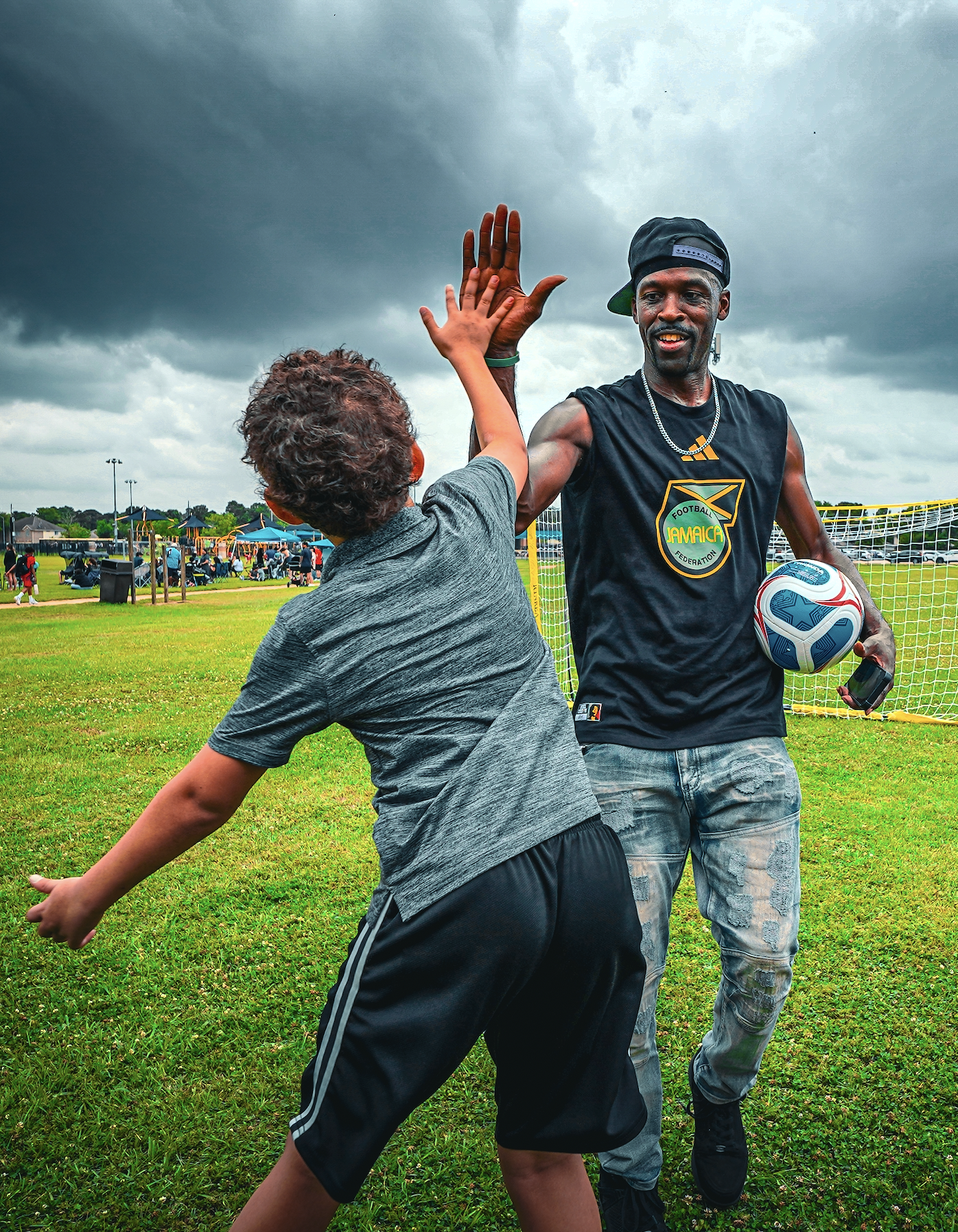 A man and a boy engaging in a playful sports moment on a grassy field, with dark clouds overhead. The man, wearing a black sleeveless shirt with a Jamaica football federation logo, jeans, and a backwards cap, is holding a football. The boy, in athletic clothing, appears to be running or chasing him. There are people and tents in the background at a park or sports field.