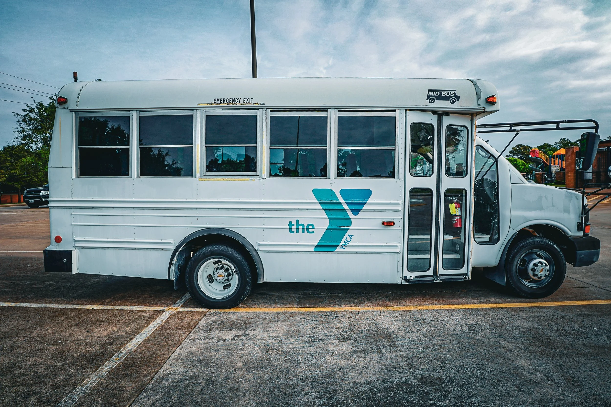 A white shuttle bus with the YMCA logo parked in an empty parking lot on a cloudy day.
