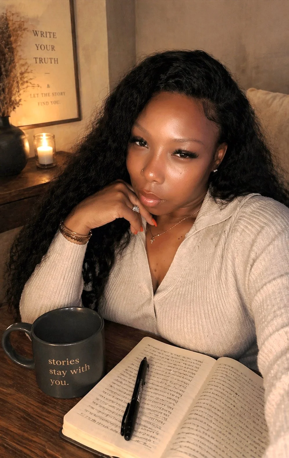 A woman with long curly hair, wearing a beige sweater, seated at a wooden table with an open book, a black pen, and a dark mug that reads 'stories stay with you.' She is resting her chin on her hand and looking at the camera. The background features a lit candle, a framed quote that says 'Write your truth - & let the story find you,' and some decorative items.