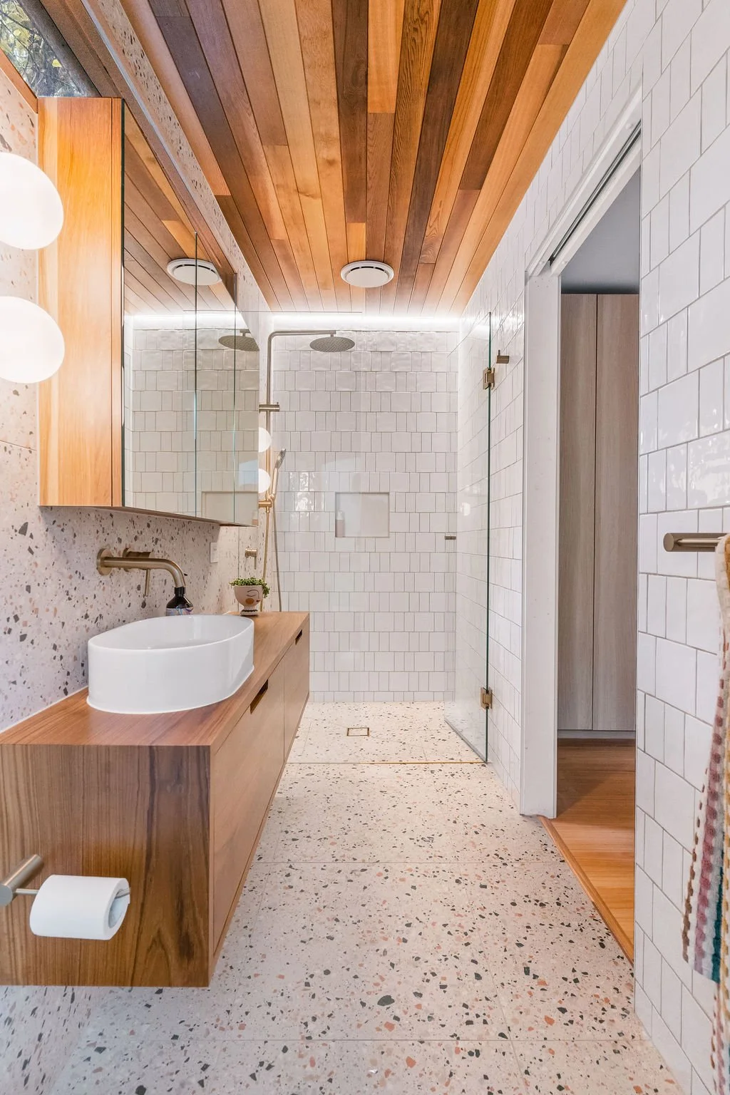 Modern bathroom with wood-paneled ceiling, white tiled shower area with glass doors, terrazzo floor, and wooden vanity with white vessel sink.