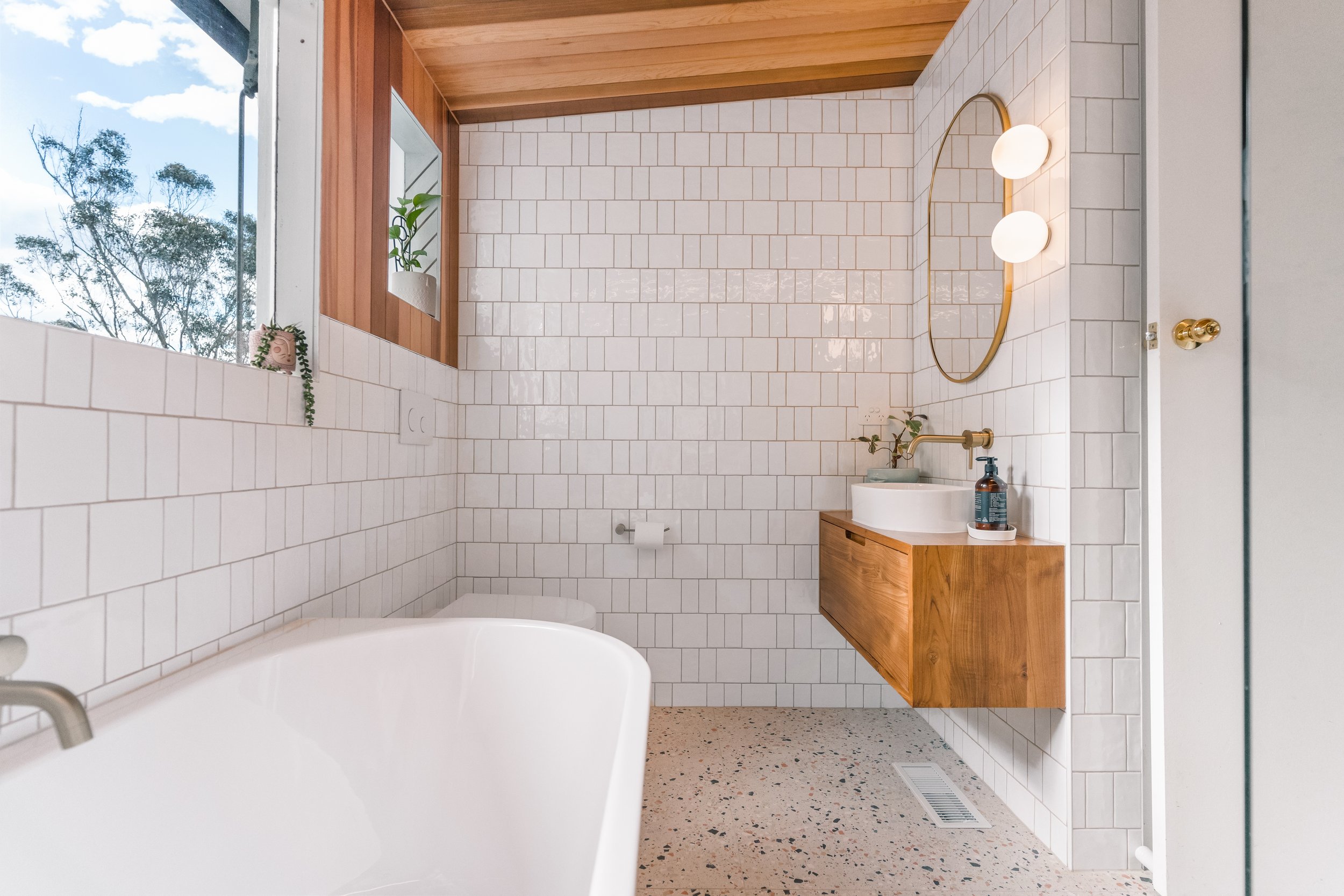 A modern bathroom with a white bathtub, white subway tile walls, a wooden vanity with a sink, frosted glass window in Yarra Valley built by Leisure Built
