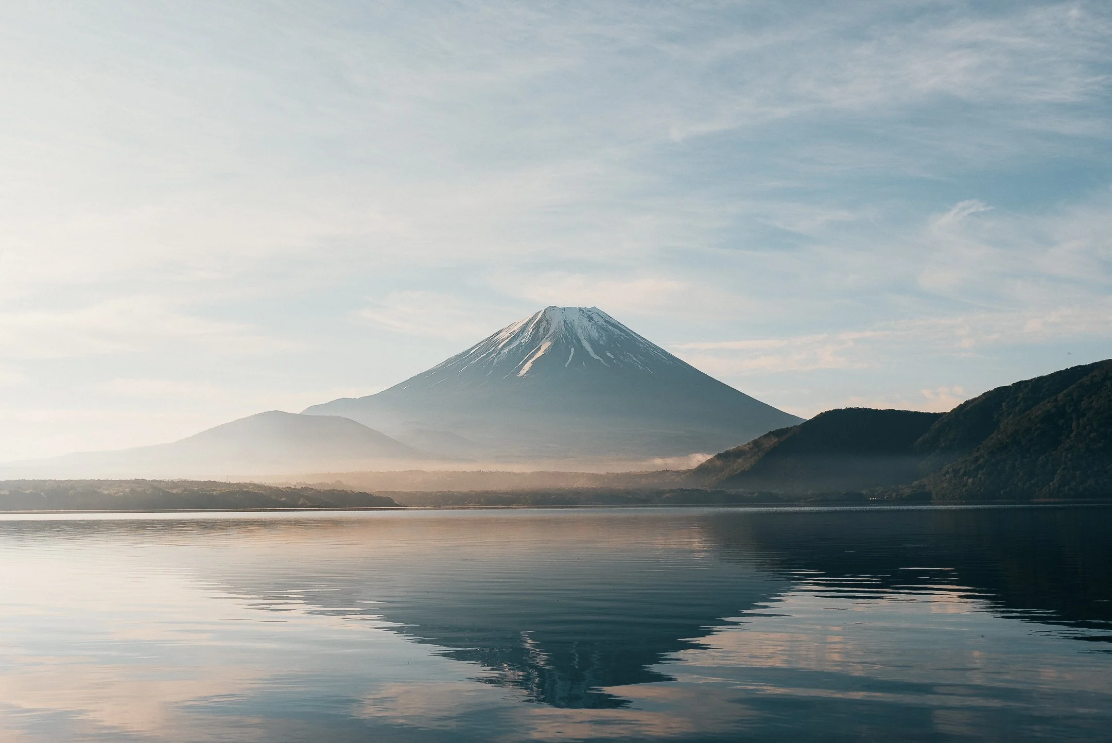 Snow-capped mountain, possibly Mount Fuji, with a lake or river in the foreground and a partly cloudy sky.