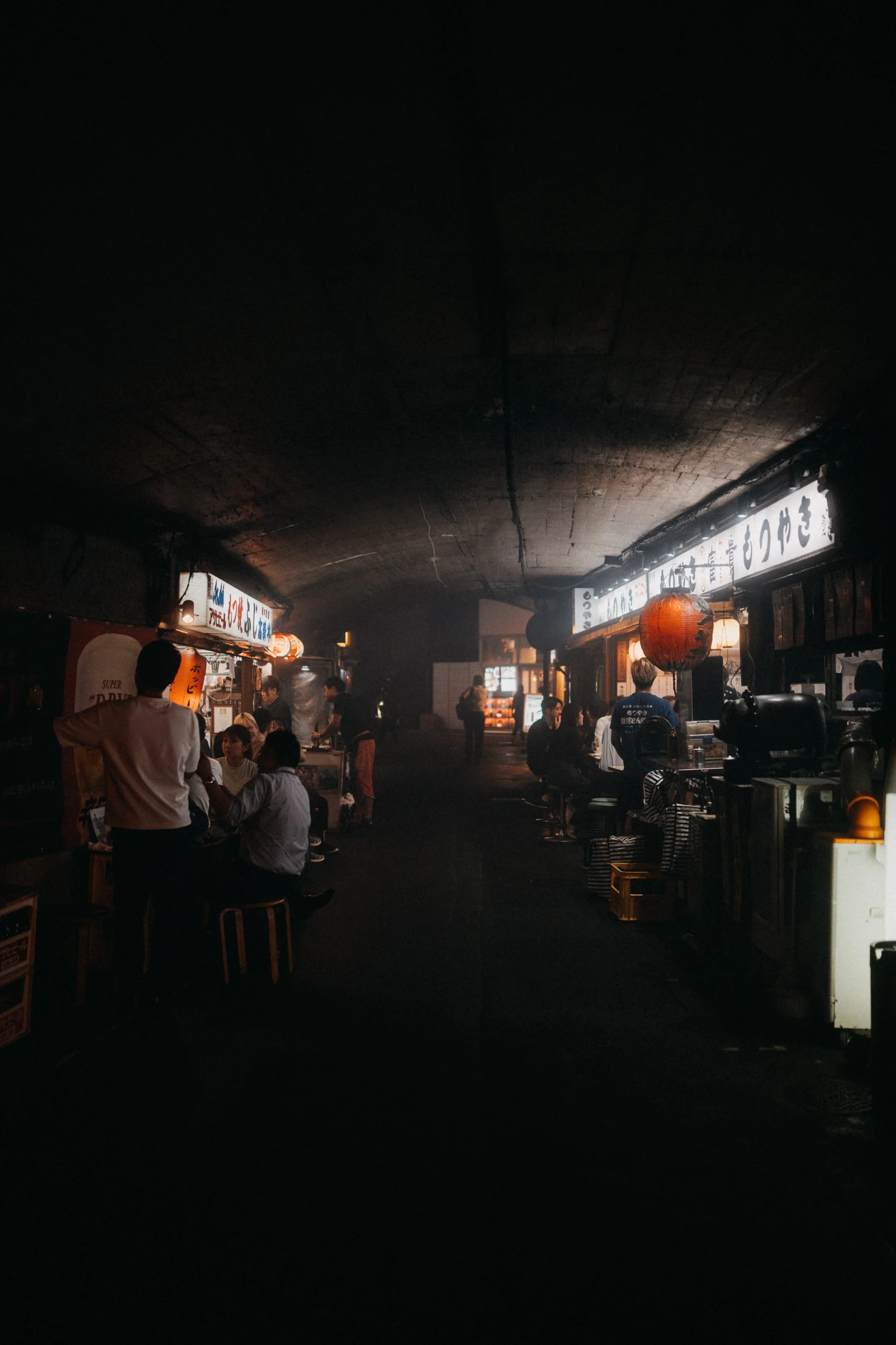A bustling Japanese restaurant or izakaya with dim lighting, Japanese signage, red paper lanterns, and people dining and socializing.
