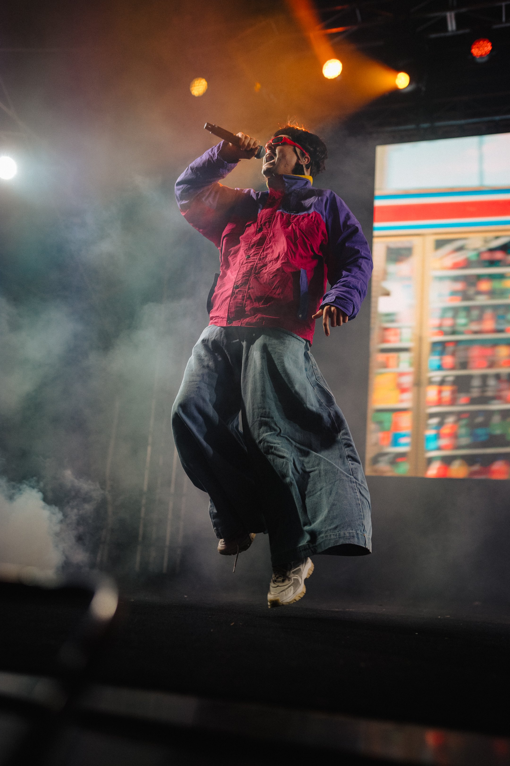 A person performing on stage with colorful lighting and smoke, wearing a red and purple jacket, jeans, red sunglasses, holding a microphone, and jumping.
