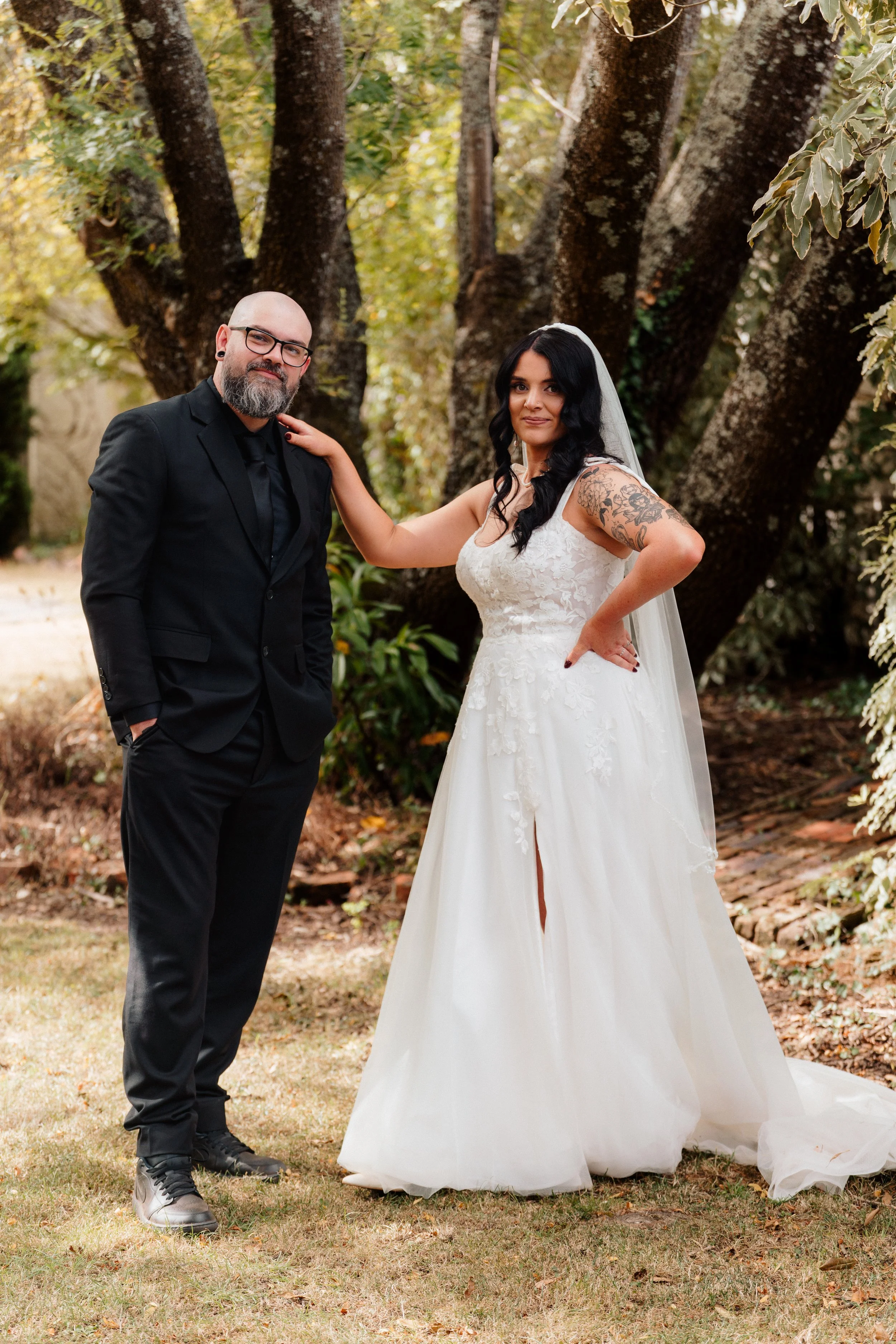 A man and woman in wedding attire standing outdoors in front of large trees, with the woman resting her hand on the man's shoulder.