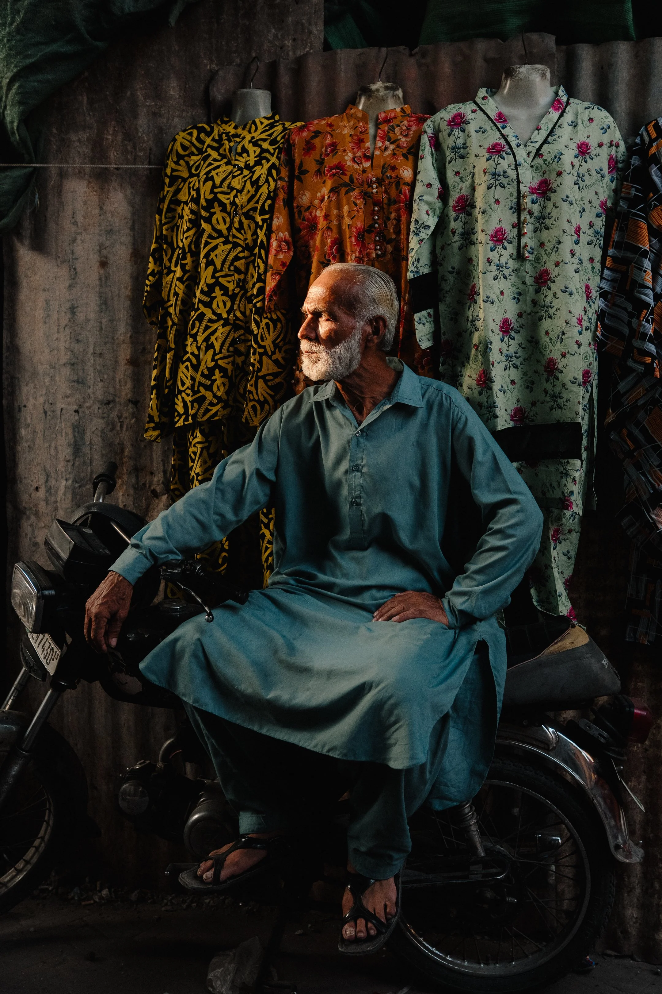 An elderly man with a white beard sitting on a motorcycle, wearing a traditional long blue kurta and black flip-flops, with three mannequins dressed in colorful floral shirts in the background.