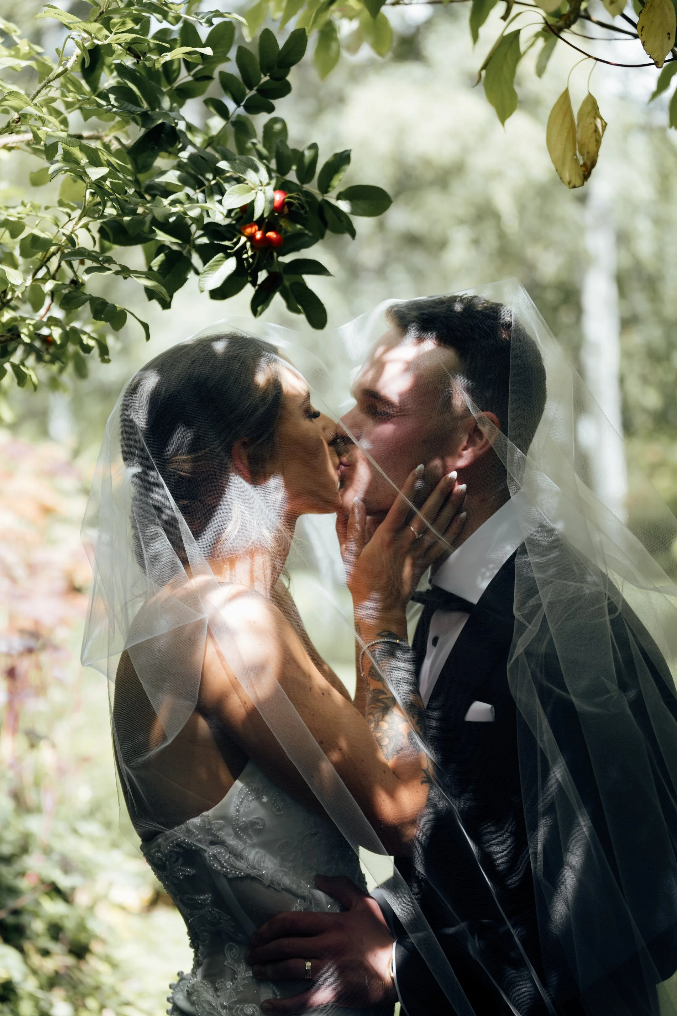 A couple sharing a tender moment and kissing outdoors under a tree, with the woman wearing a wedding dress and veil, and the man in a tuxedo, both wrapped in a sheer veil, surrounded by green foliage.