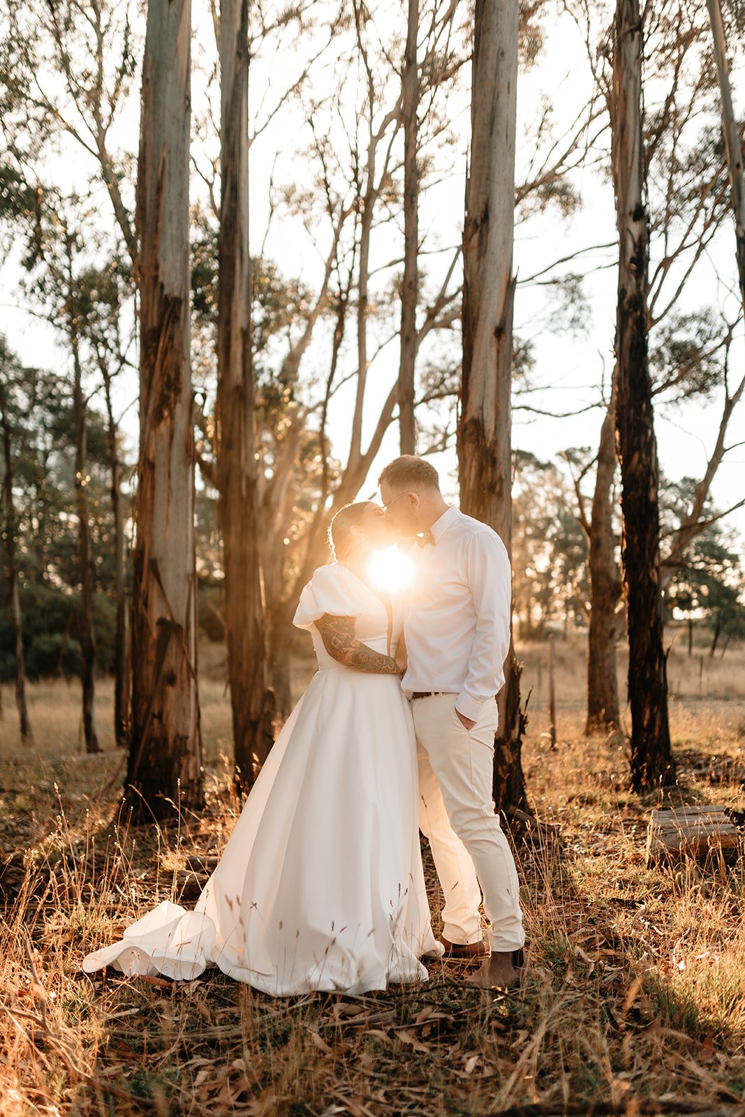 A couple in wedding attire kissing in a forest at sunset, with the sun shining between them.