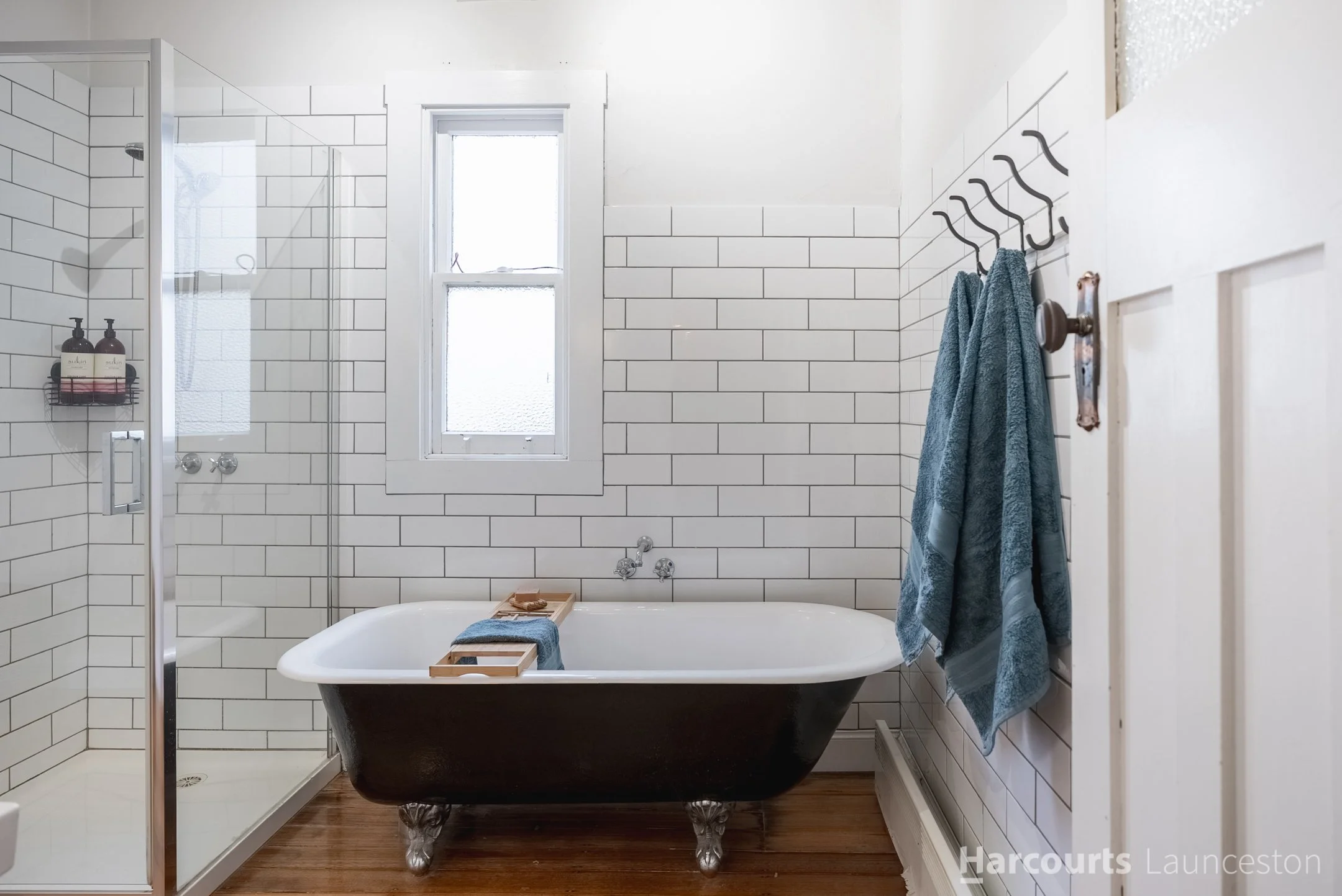 Bathroom with white subway tile walls and a clawfoot bathtub. A window above the tub, a towel hanging on a wall hook, and a shower enclosure are visible.