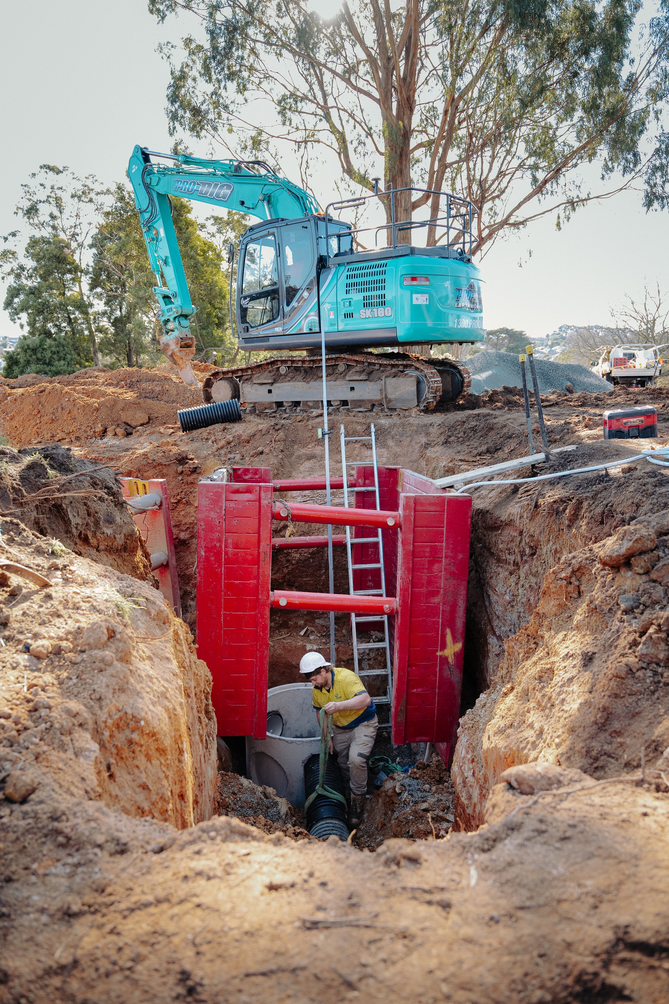 Construction site with an excavator on the ground and a worker installing piping in a trench surrounded by dirt and trees.