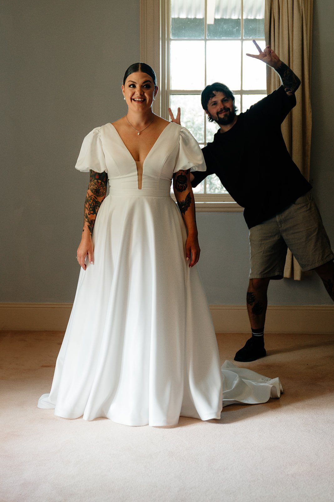 A woman in a wedding dress standing indoors with a man making a peace sign behind her near a window.