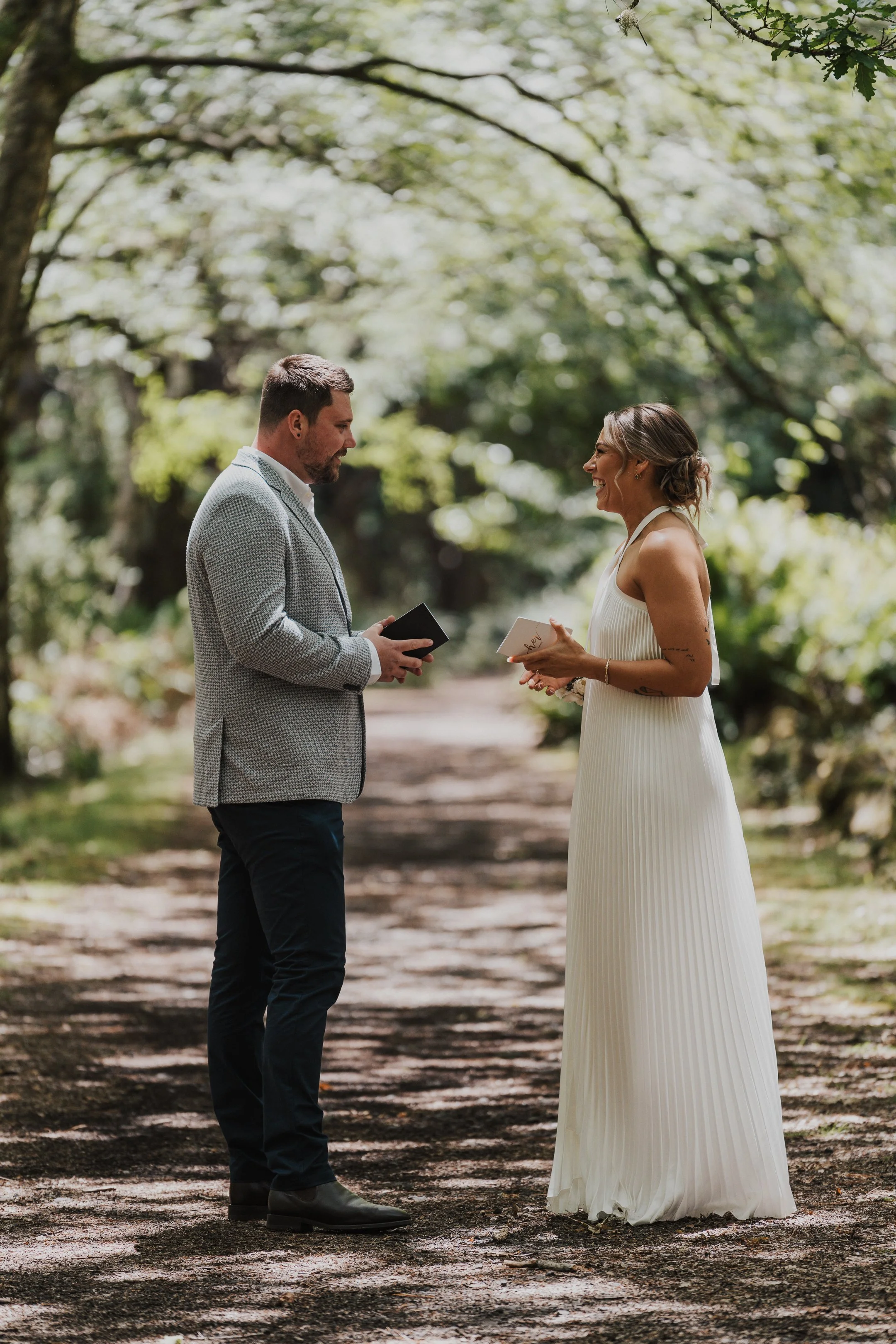 A man and woman exchange vows in a wooded outdoor setting, both holding small books, with strings of light and greenery overhead.