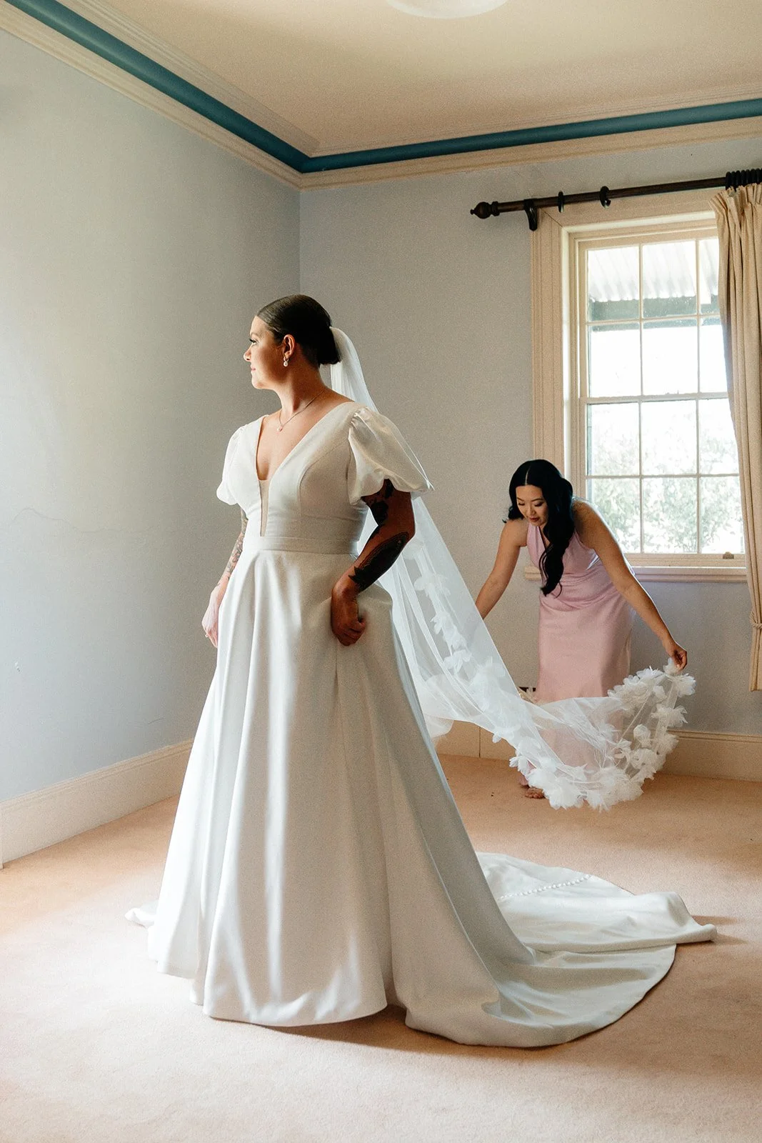 A bride in a white wedding dress standing in a room, with a bridesmaid in a pink dress adjusting the train of the dress.