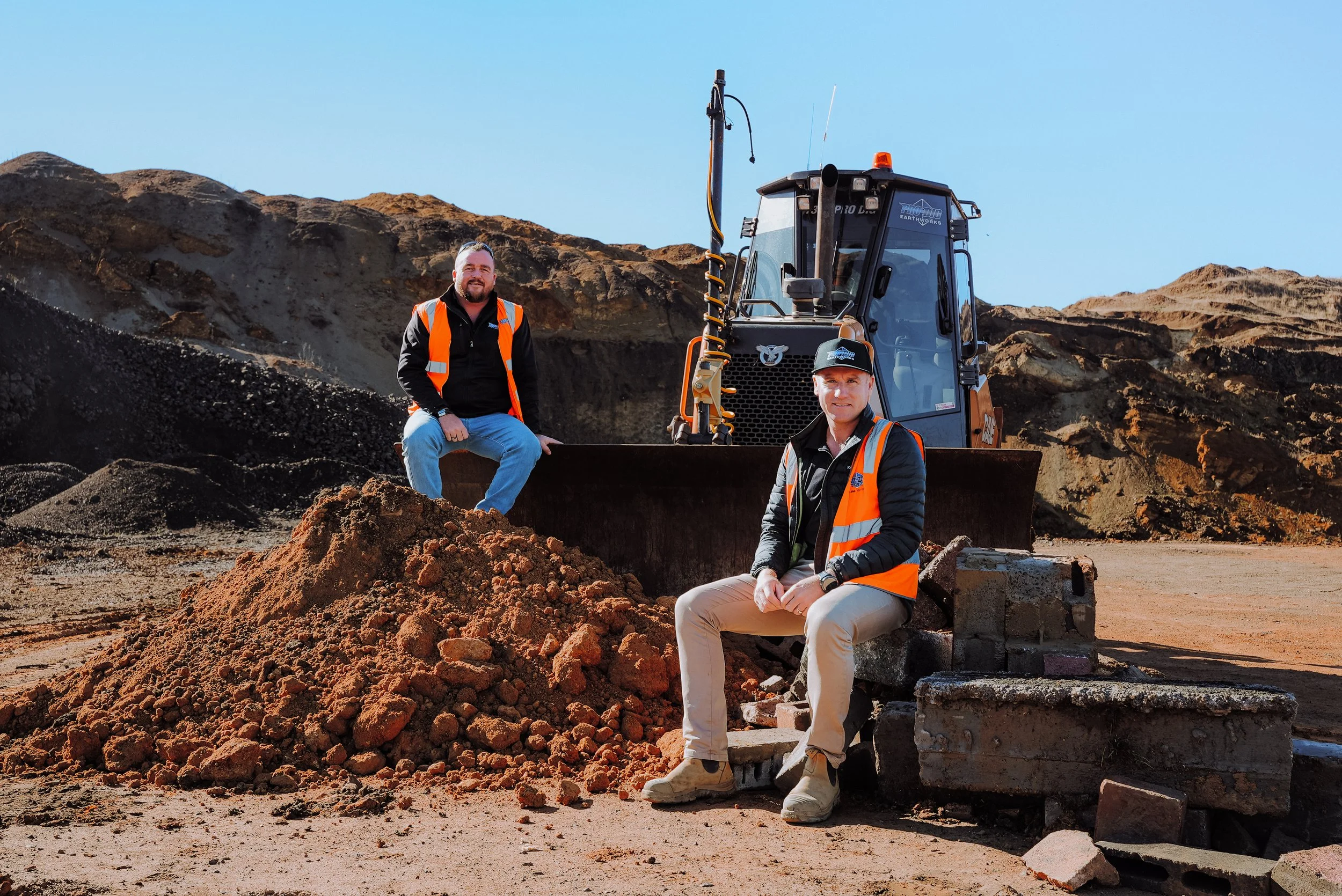 Two men in orange safety vests sitting on and near a construction site with dirt and soil mounds, a bulldozer in the background, under a clear blue sky.