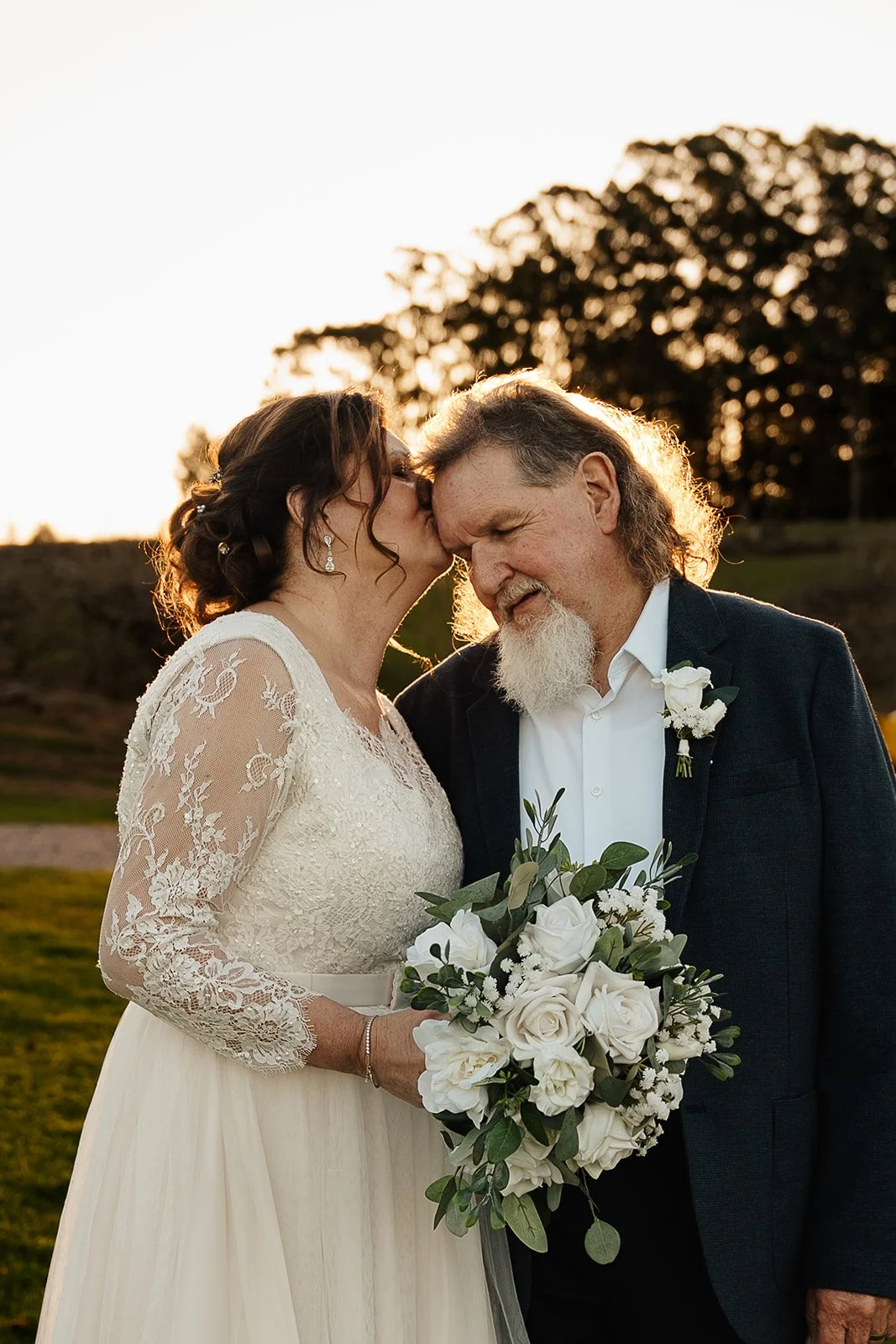 A bride and an older man, likely her father, sharing a tender moment during a wedding ceremony at sunset, with trees in the background. The bride is wearing a lace wedding dress and holding a bouquet of white roses and greenery.