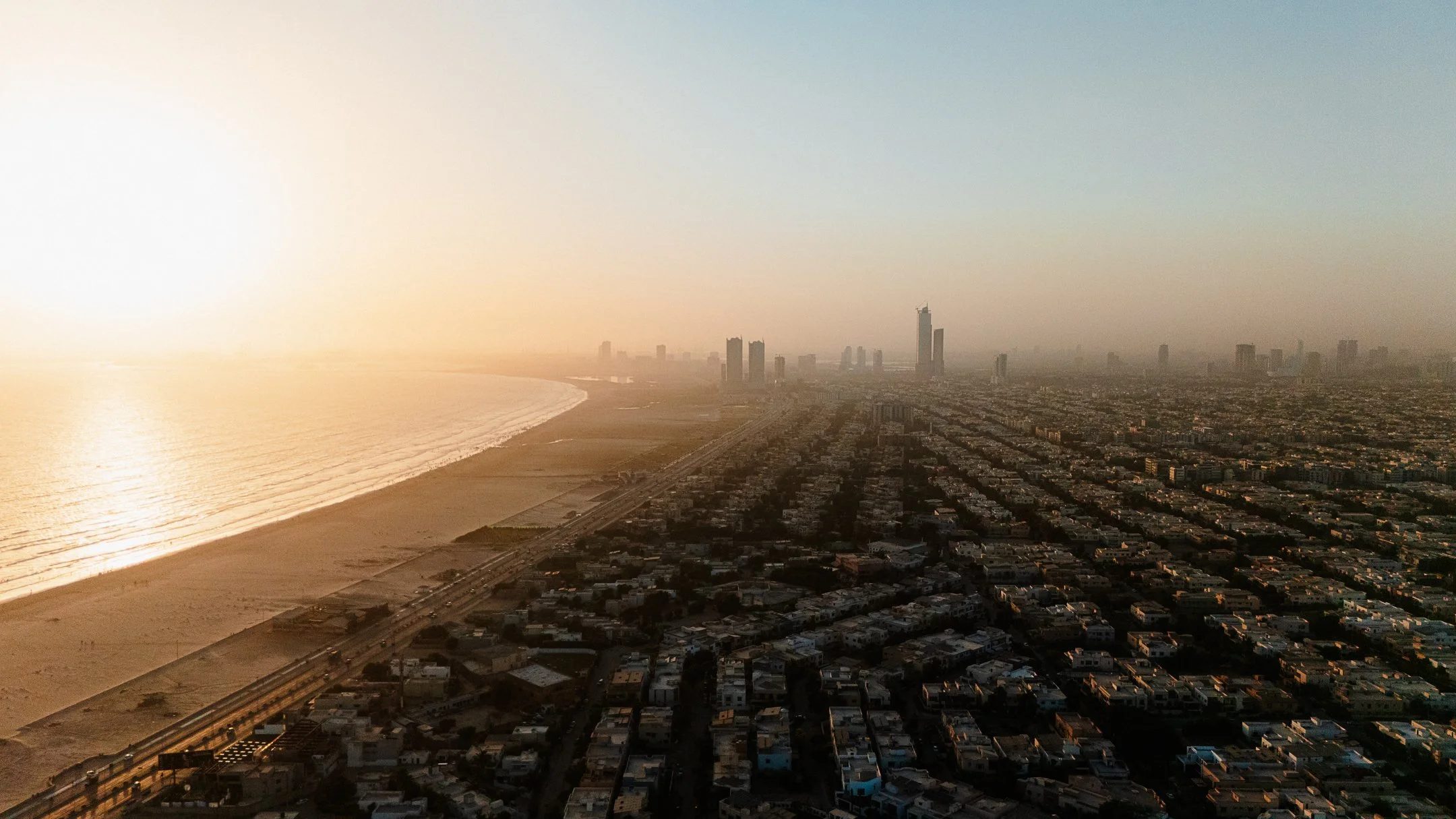 Aerial view of a cityscape with high-rise buildings along the coastline at sunset, with the beach and ocean in the foreground.