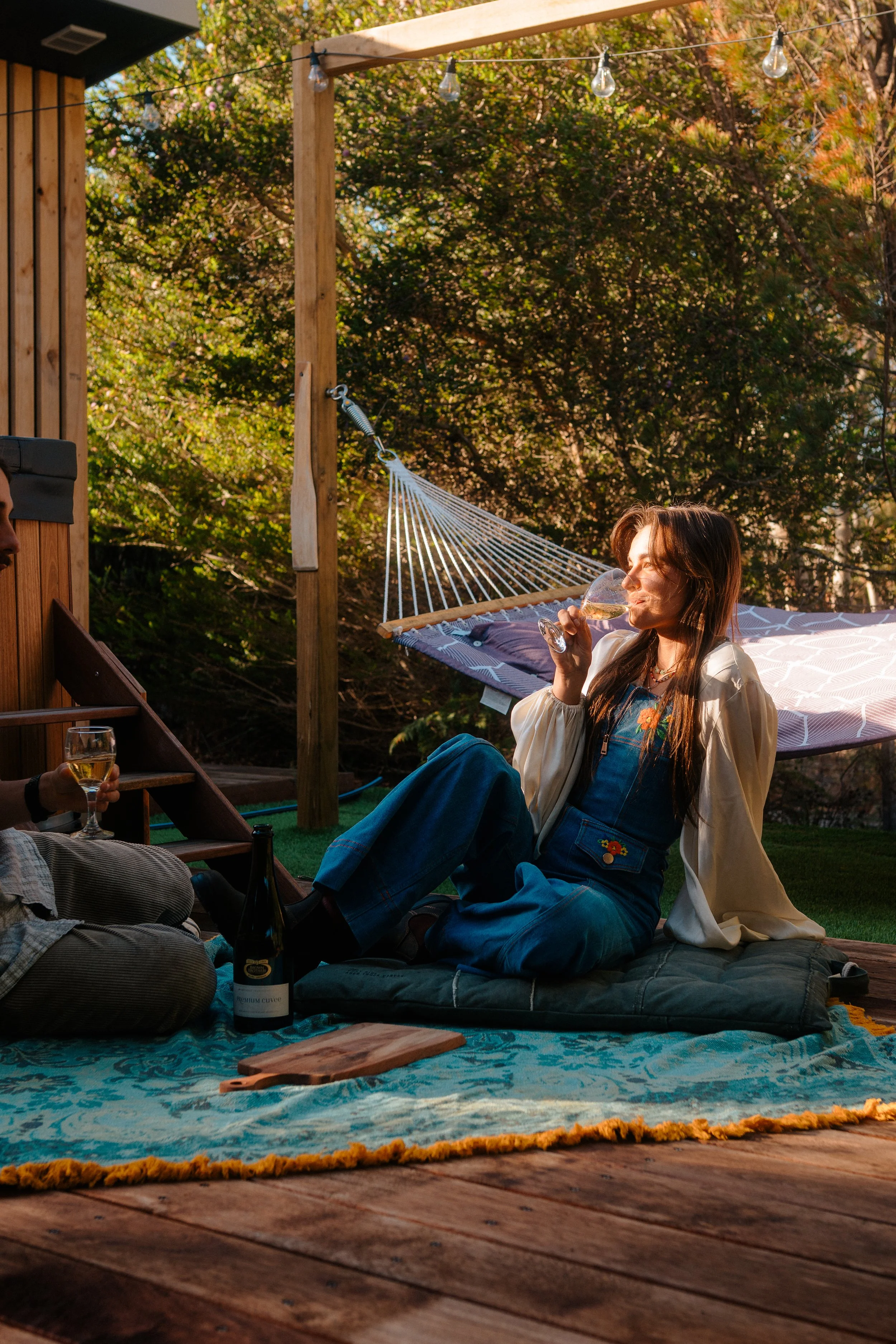 A woman sitting outside on a blanket, drinking wine, with a hammock and trees in the background, during daytime.
