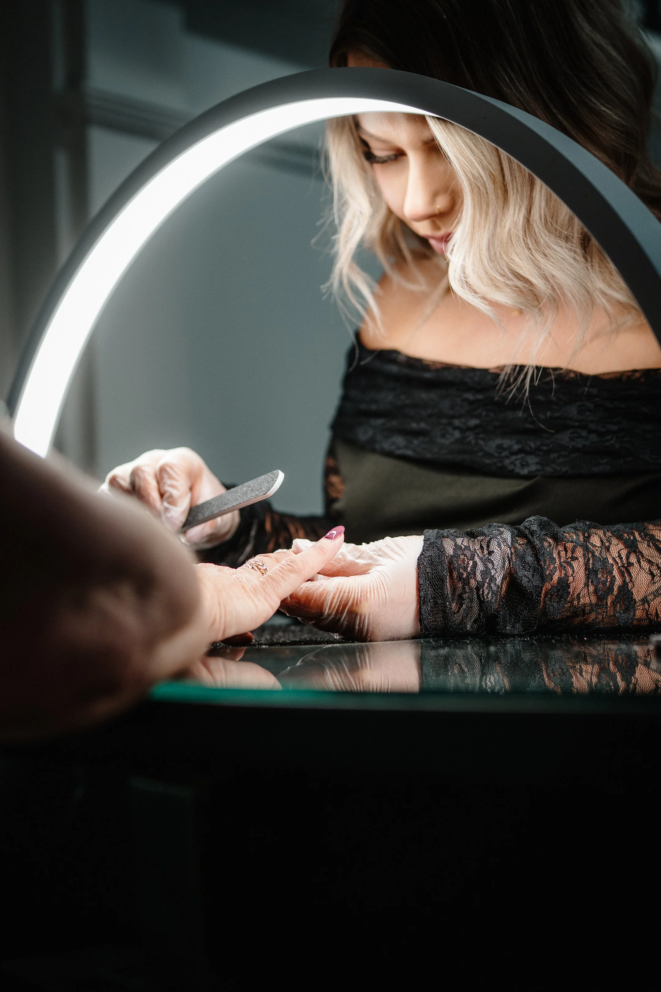 A woman with blonde hair appears to be receiving a manicure at a salon. She is wearing a black lace top and looking down as a nail technician, who is wearing gloves, works on her nails. An arc-shaped light illuminates the scene.