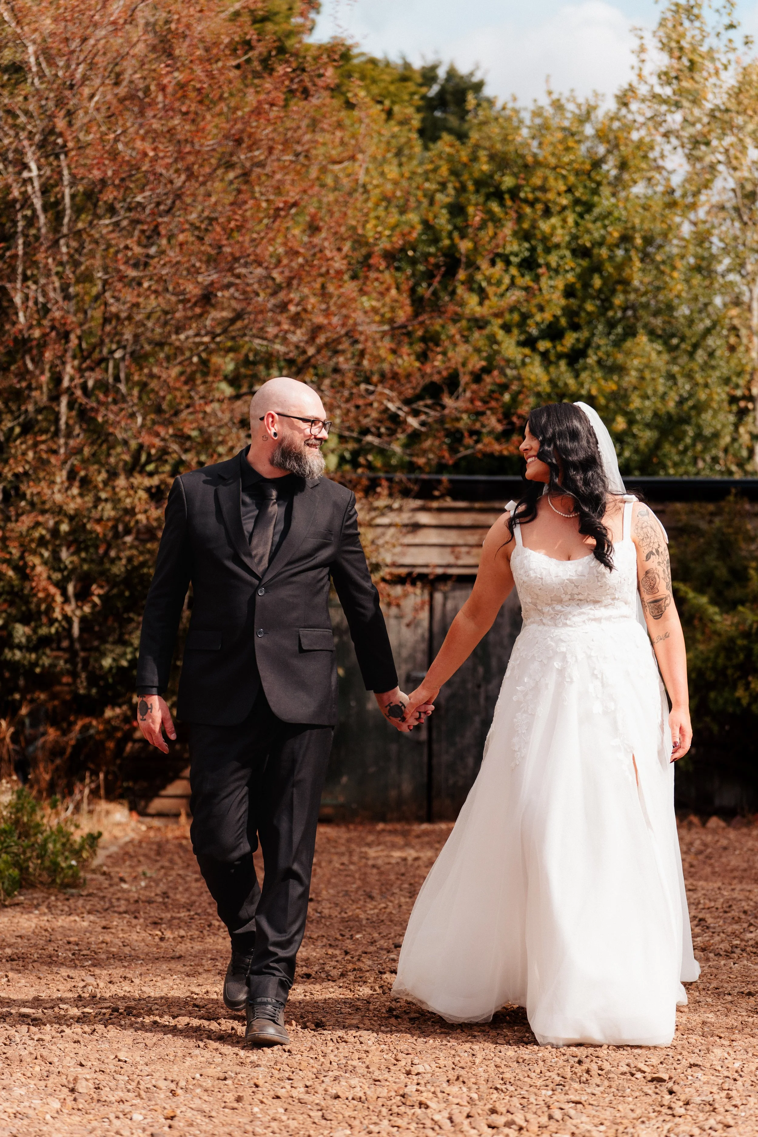 A couple dressed in wedding attire holding hands outdoors on a fall day, with trees in autumn colors in the background.