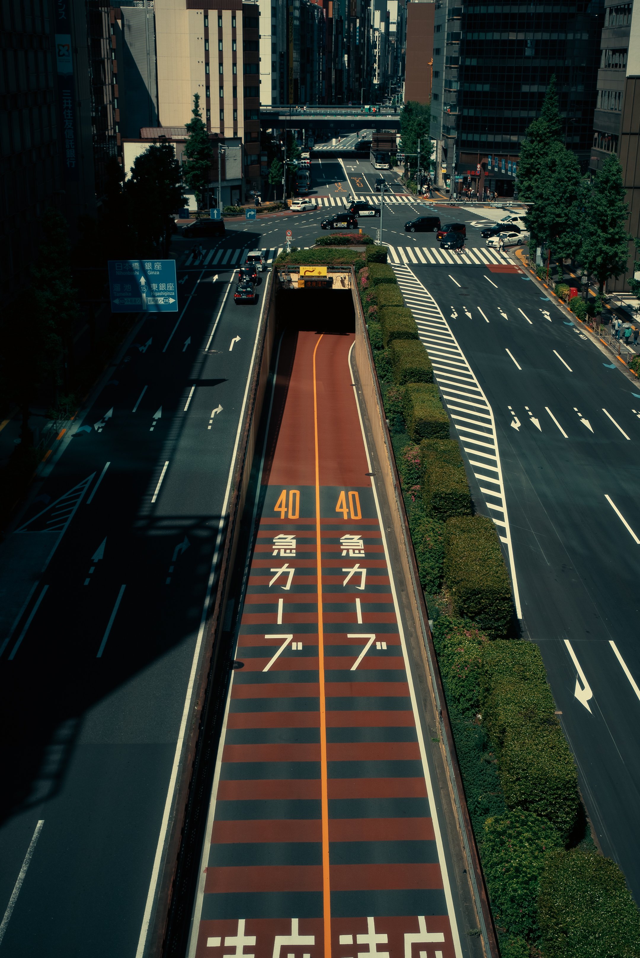 A busy city street intersecting with multiple lanes and roads, with several cars, trees lining the sidewalks, and tall buildings in the background. There are traffic signs and pedestrian crosswalks visible.