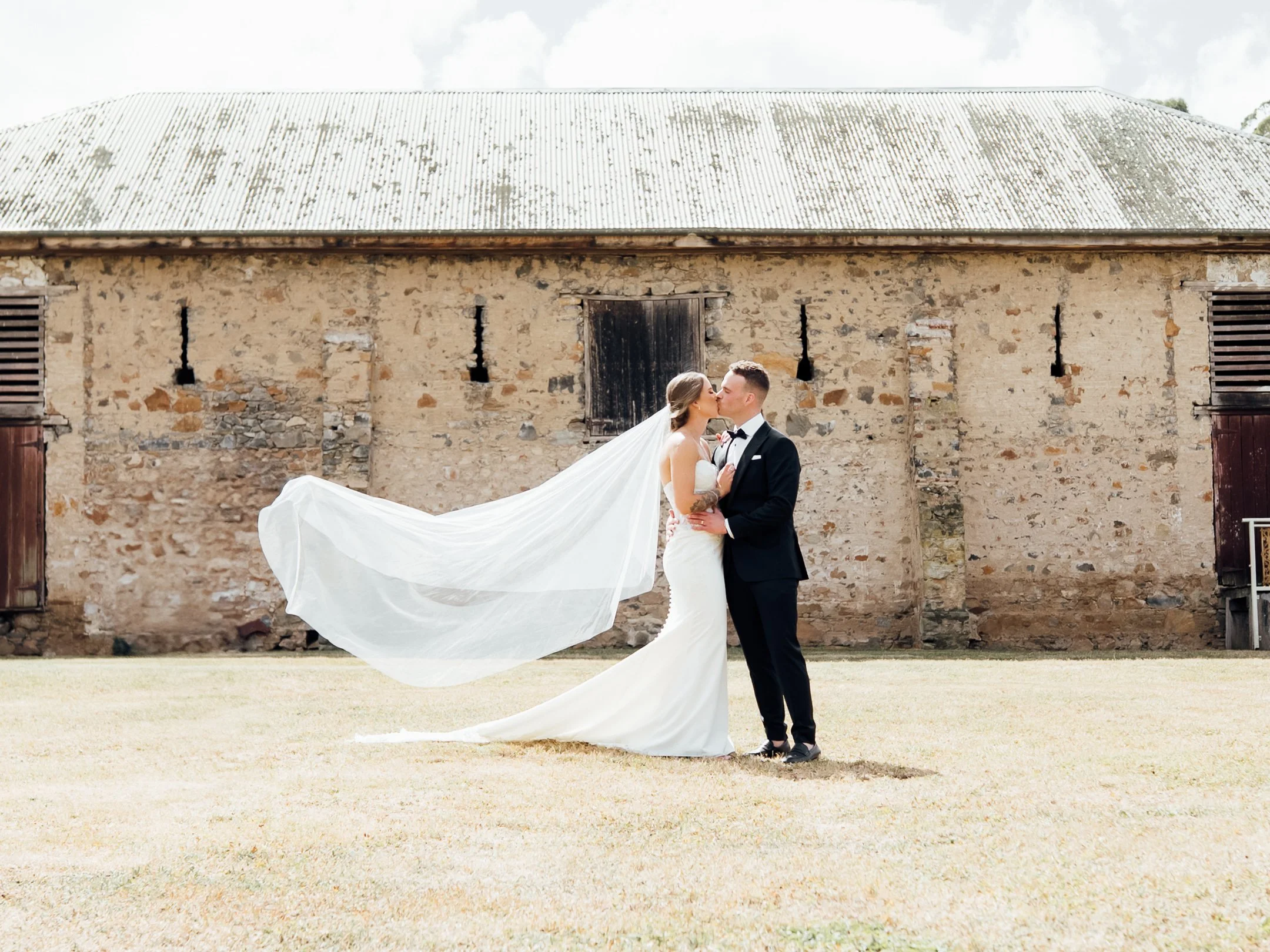 A bride and groom share a kiss outdoors in front of an old stone building, with the bride wearing a white wedding gown and veil, and the groom in a black tuxedo.