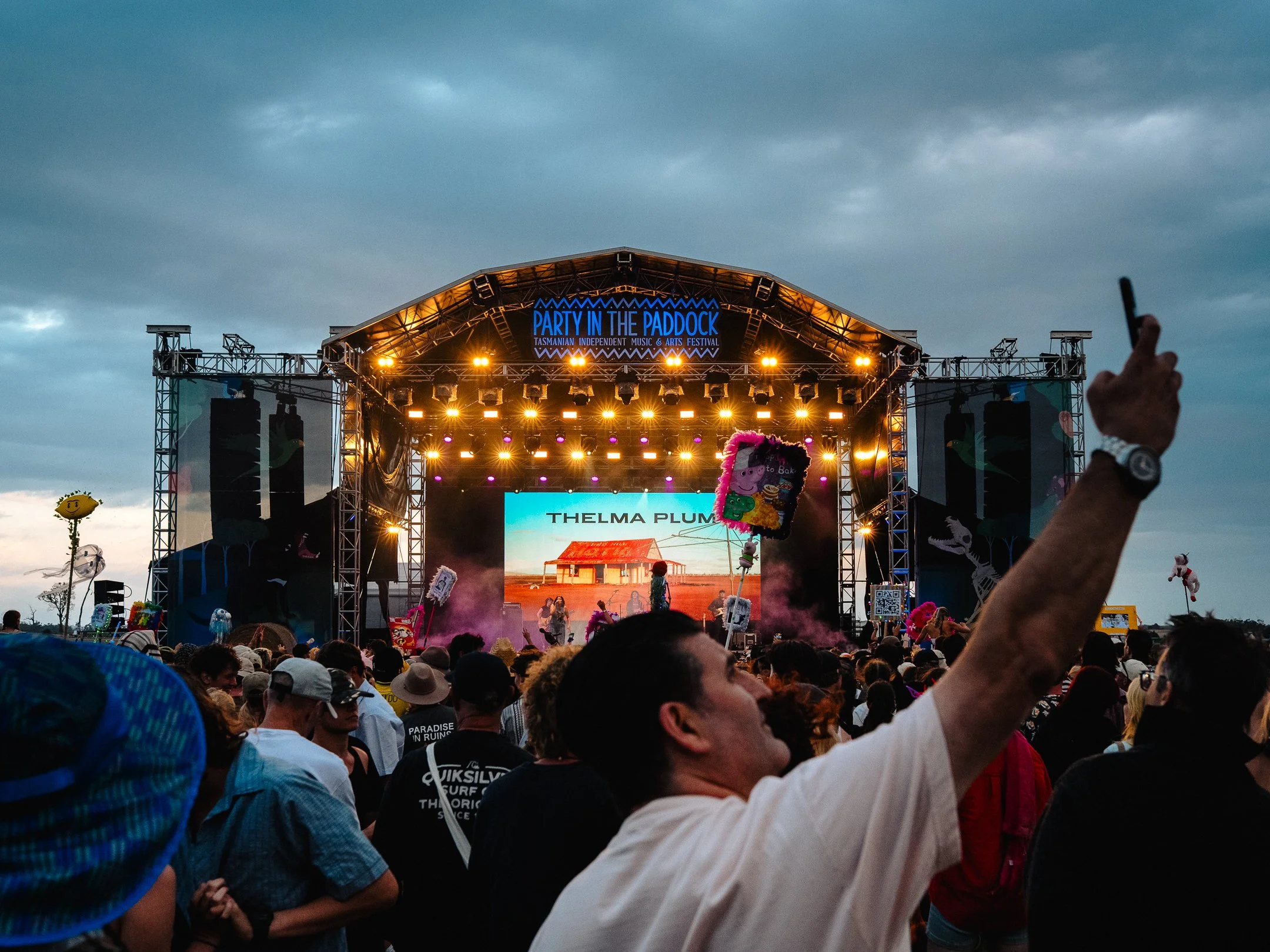 Crowd of people attending an outdoor music festival with a large stage displaying the sign 'Party in the Paddock' and a screen showing the name 'Thelma Plum.' The sky is overcast, and the audience is enjoying the event with some holding up signs and 