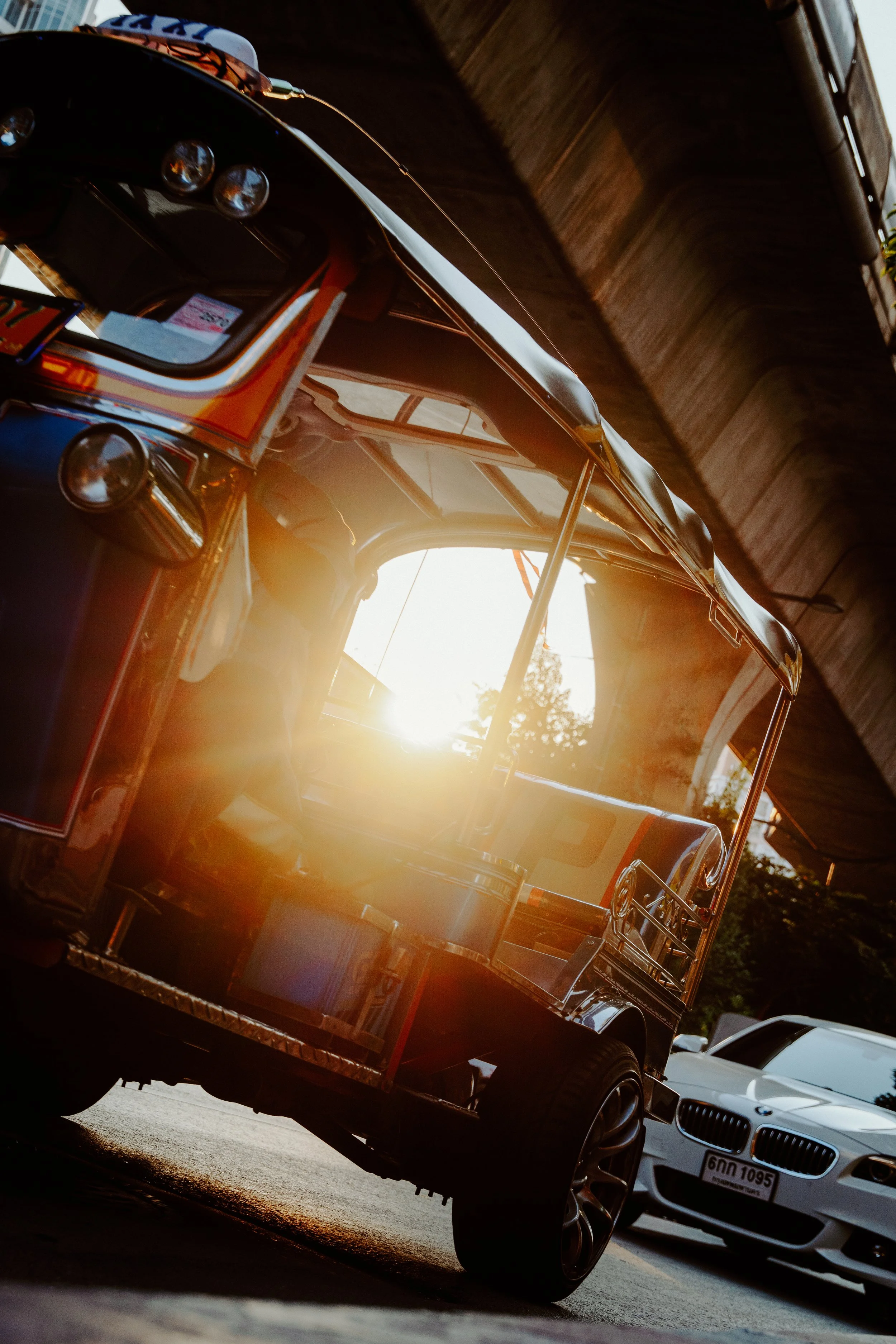 Low-angle view of a food cart with shiny metal surfaces and lights, parked on the street at sunset sunlight glinting off its chrome finish, with two white sedans parked nearby.