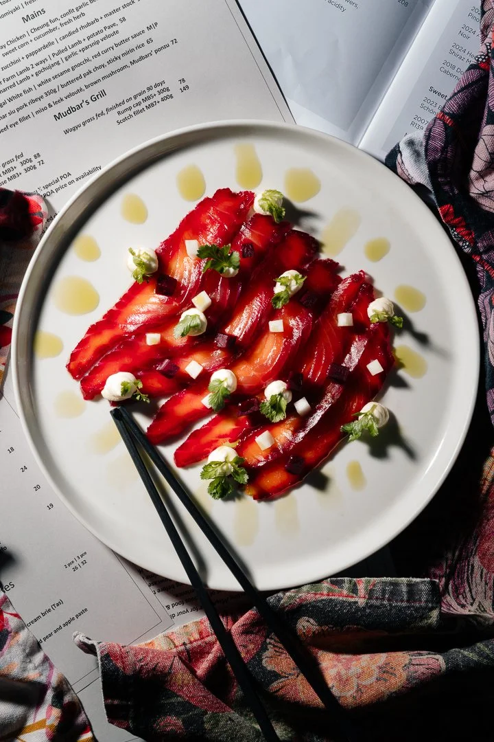 A white plate with sliced strawberries garnished with small dollops of cream or cheese and tiny green herbs, drizzled with a yellow sauce, served with black chopsticks on a table with menu pages and fabric.