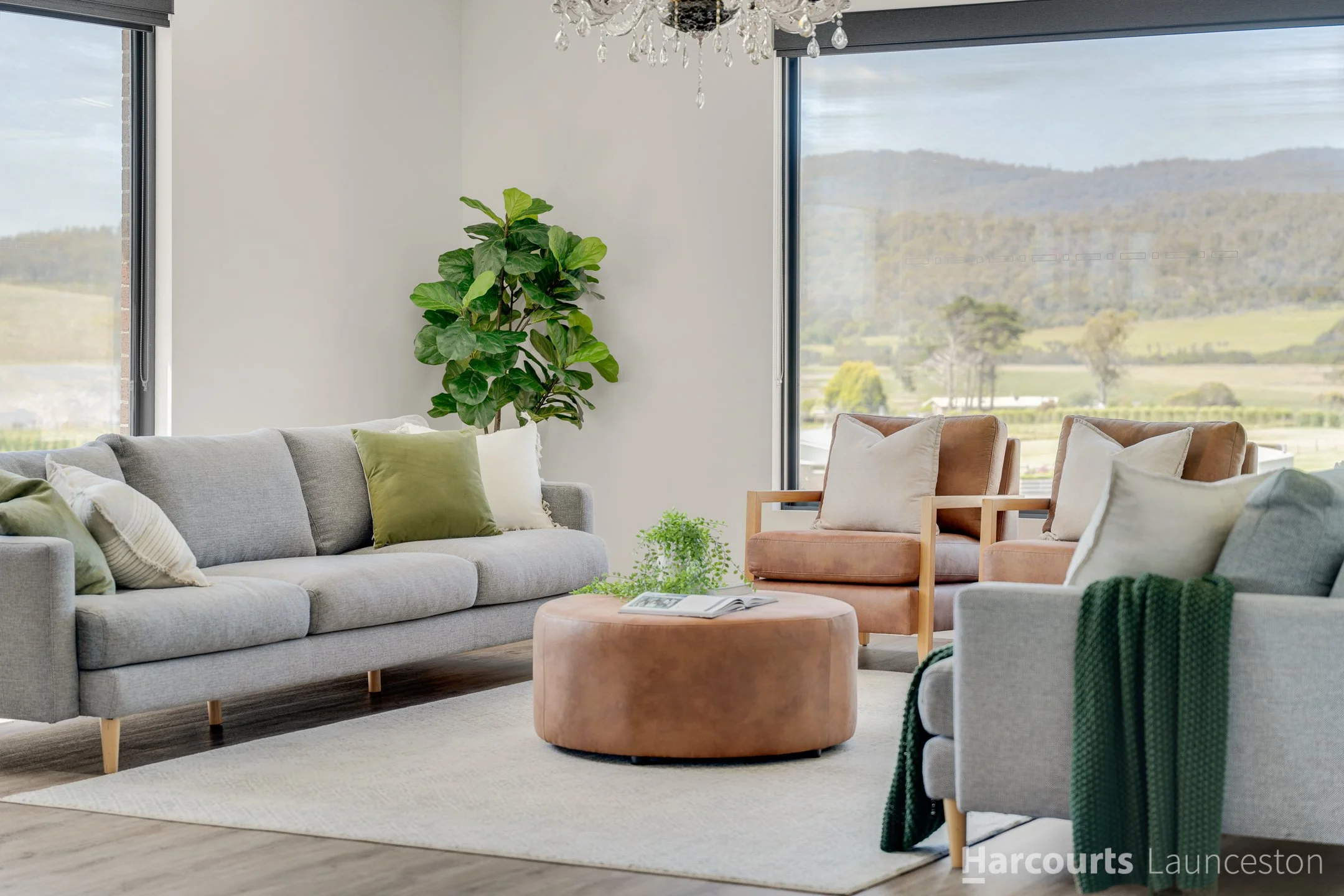 Modern living room with a large gray sofa with green and white pillows, a round pink ottoman, a beige armchair, a wooden side chair, a large green potted plant, a chandelier, and large windows showing a scenic landscape with hills and trees.