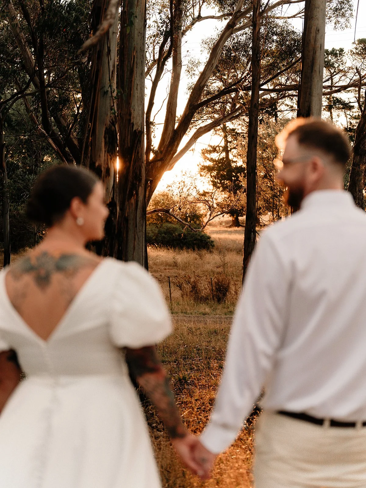 A couple holding hands during a sunset in a natural outdoor setting with trees and grass, seen from behind