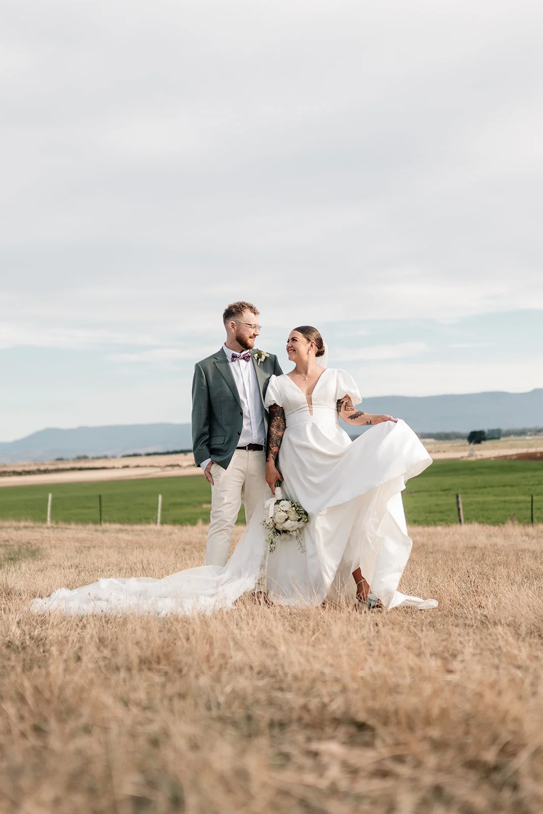 A bride and groom standing in a field, with the bride lifting her gown to show her shoes, holding a bouquet, and looking at each other, with a scenic landscape and cloudy sky in the background.