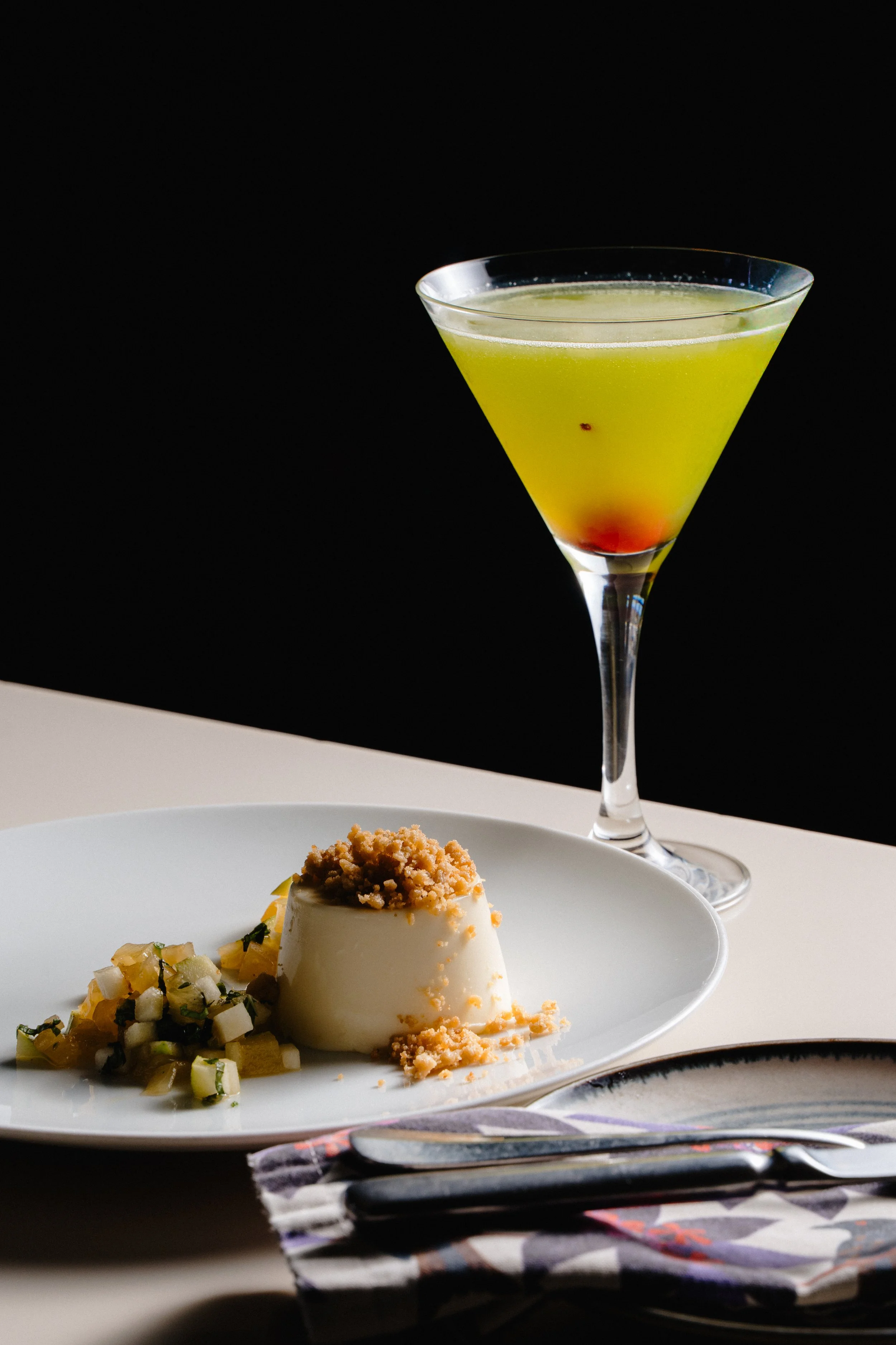 A dessert on a white plate with a scoop of ice cream topped with crumbles, accompanied by a small side of diced fruit salad, and a cocktail in a martini glass with yellow liquid and a red garnish at the bottom, set against a dark background.
