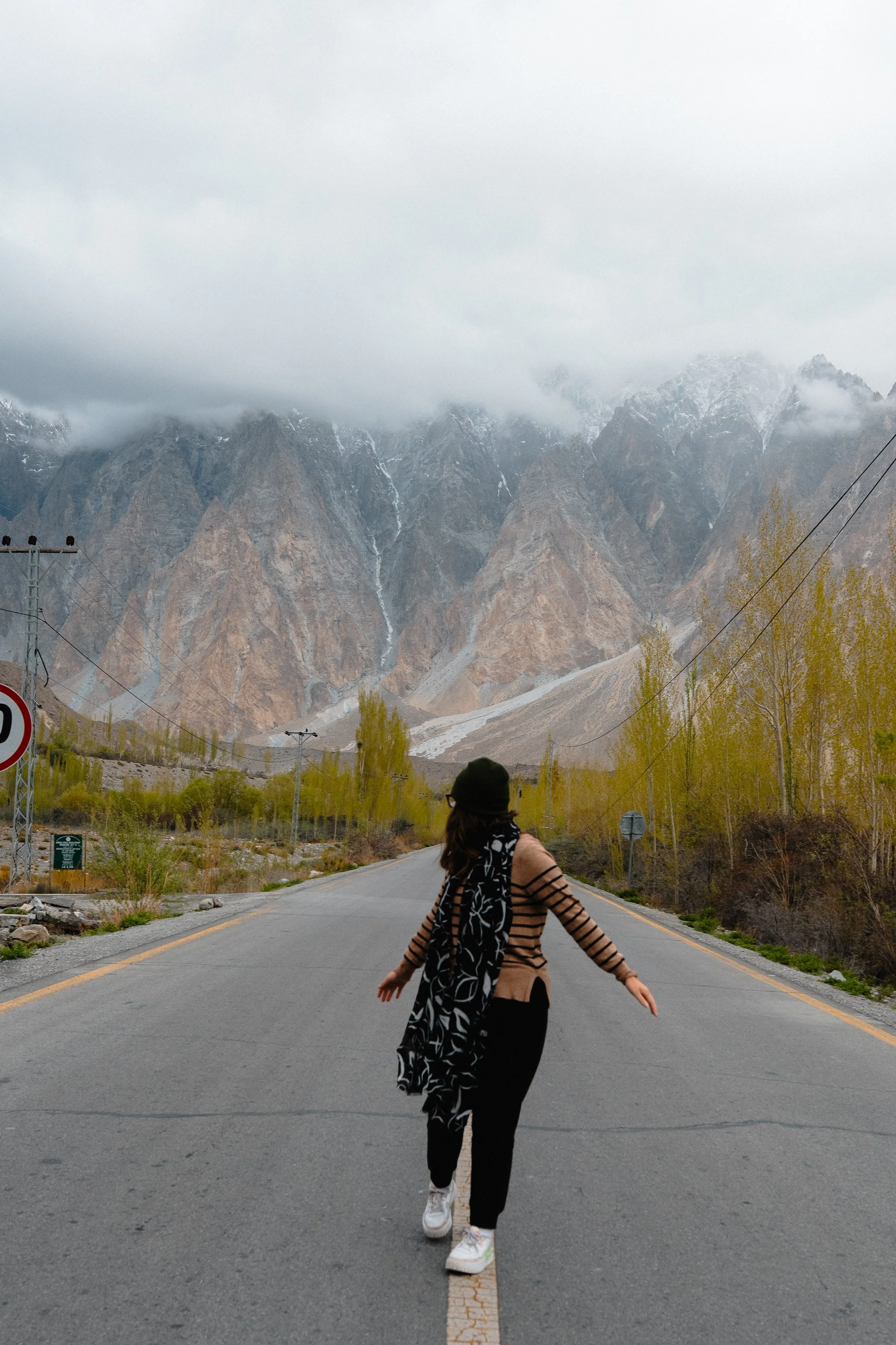 A woman walking across a mountain road with tall, rocky mountains and cloudy sky in the background.