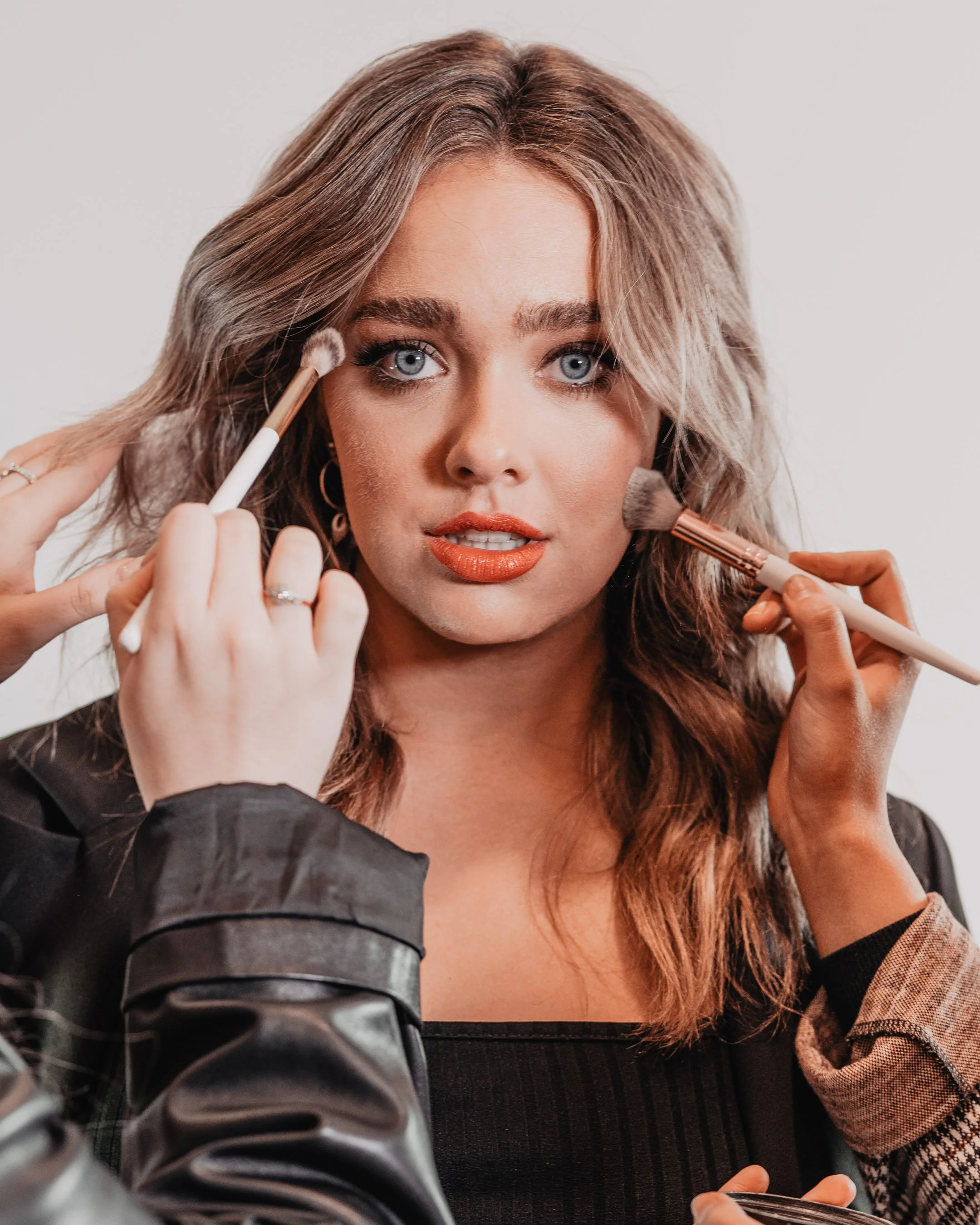 A woman with wavy light brown hair and blue eyes applying makeup with brushes, wearing black and plaid clothing.
